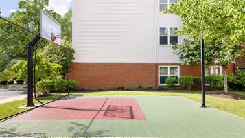 Una cancha de baloncesto con superficie roja, rodeada por un edificio de ladrillo y árboles.