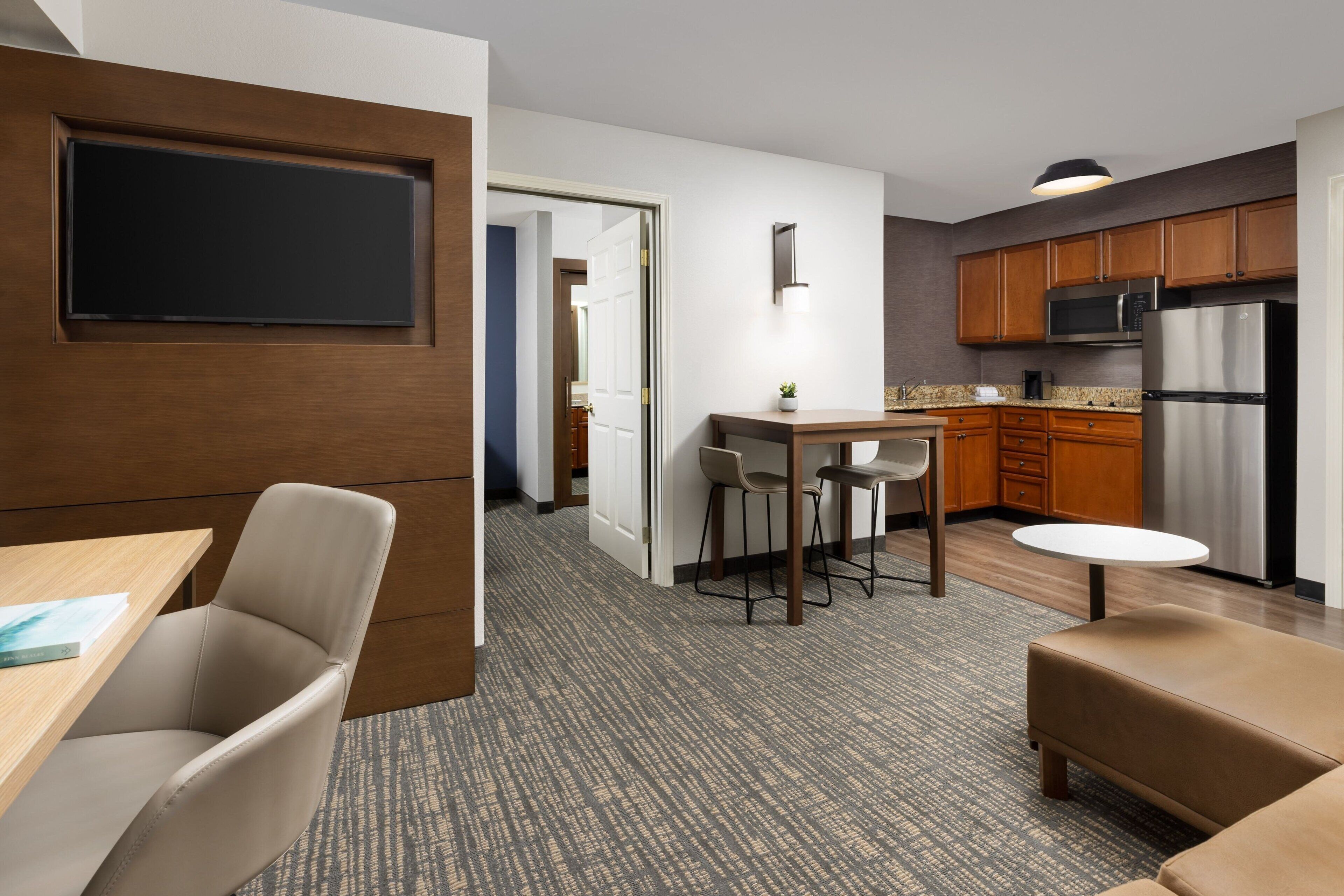 A modern hotel room with a flat-screen TV mounted on a wooden panel, a dining area with a table and chairs, a kitchenette with stainless steel appliances, and a cozy seating area with a sofa and ottoman.