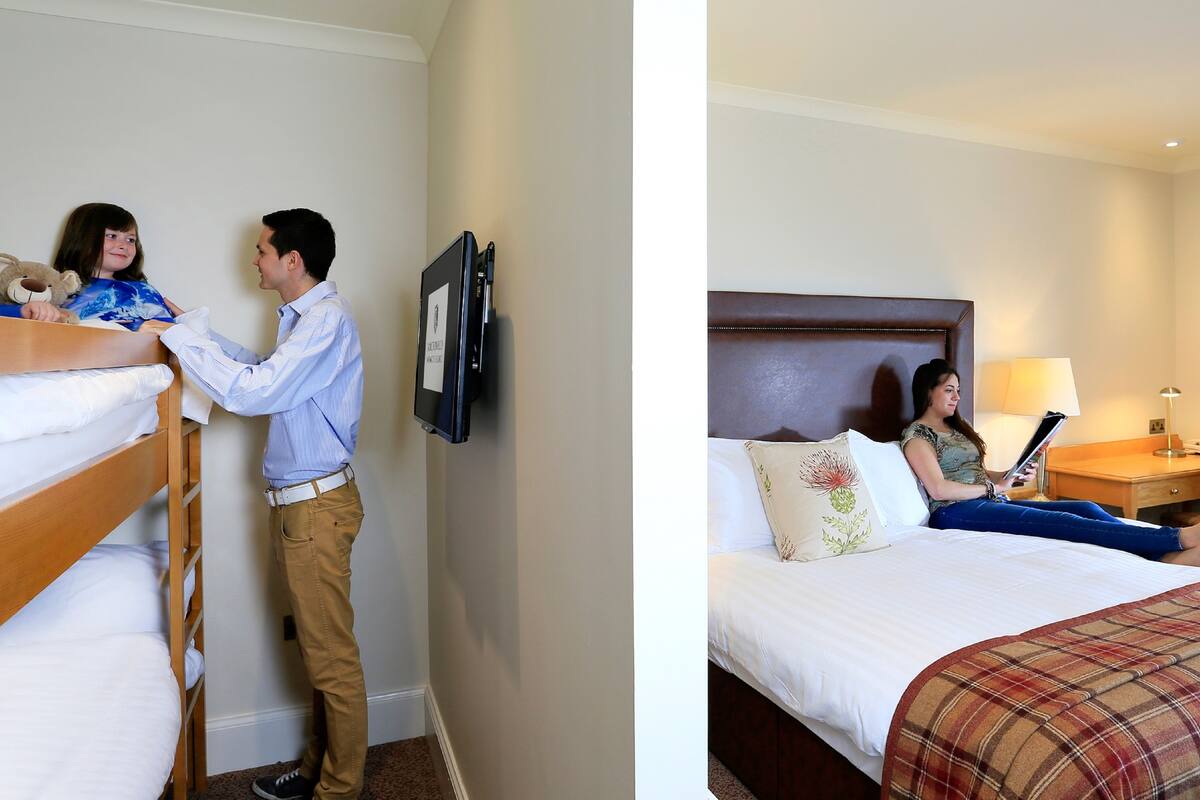 A hotel room with a bunk bed, a bedside table with a lamp, and a bed with a plaid blanket.