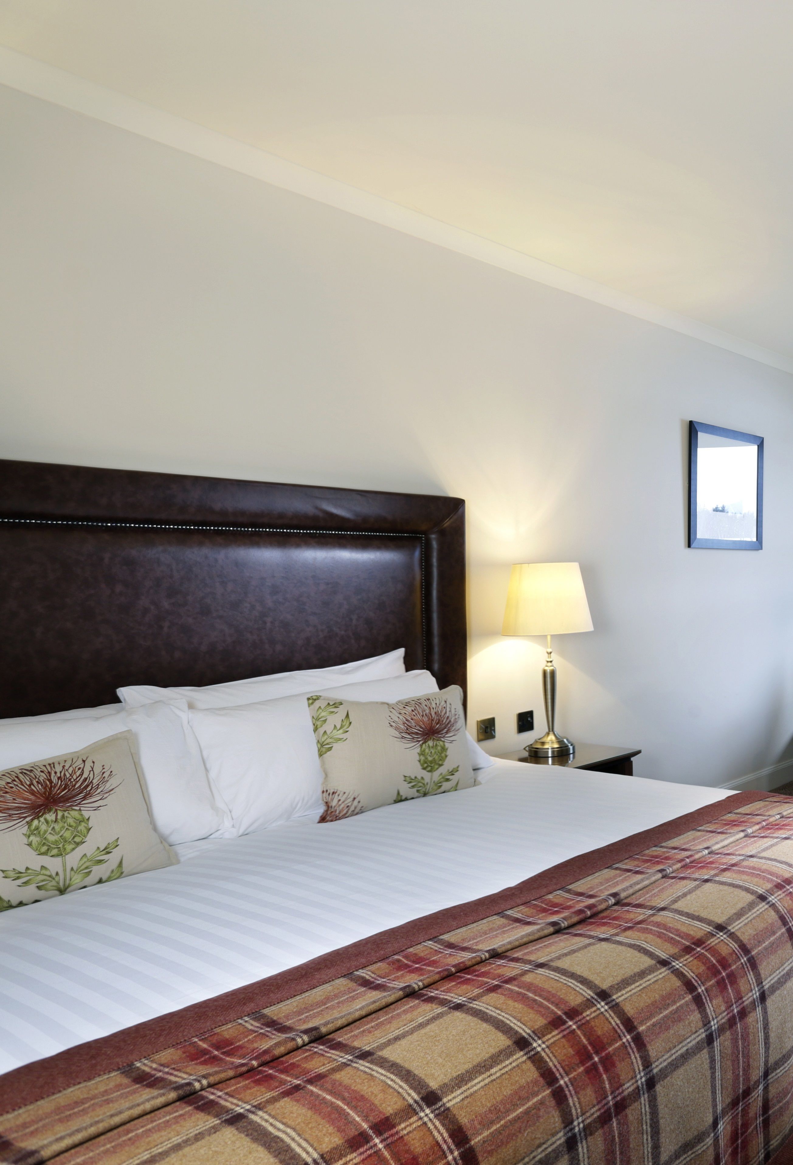 Een net opgemaakt bed met een geruit sprei, een houten hoofdbord en een bedlampje.