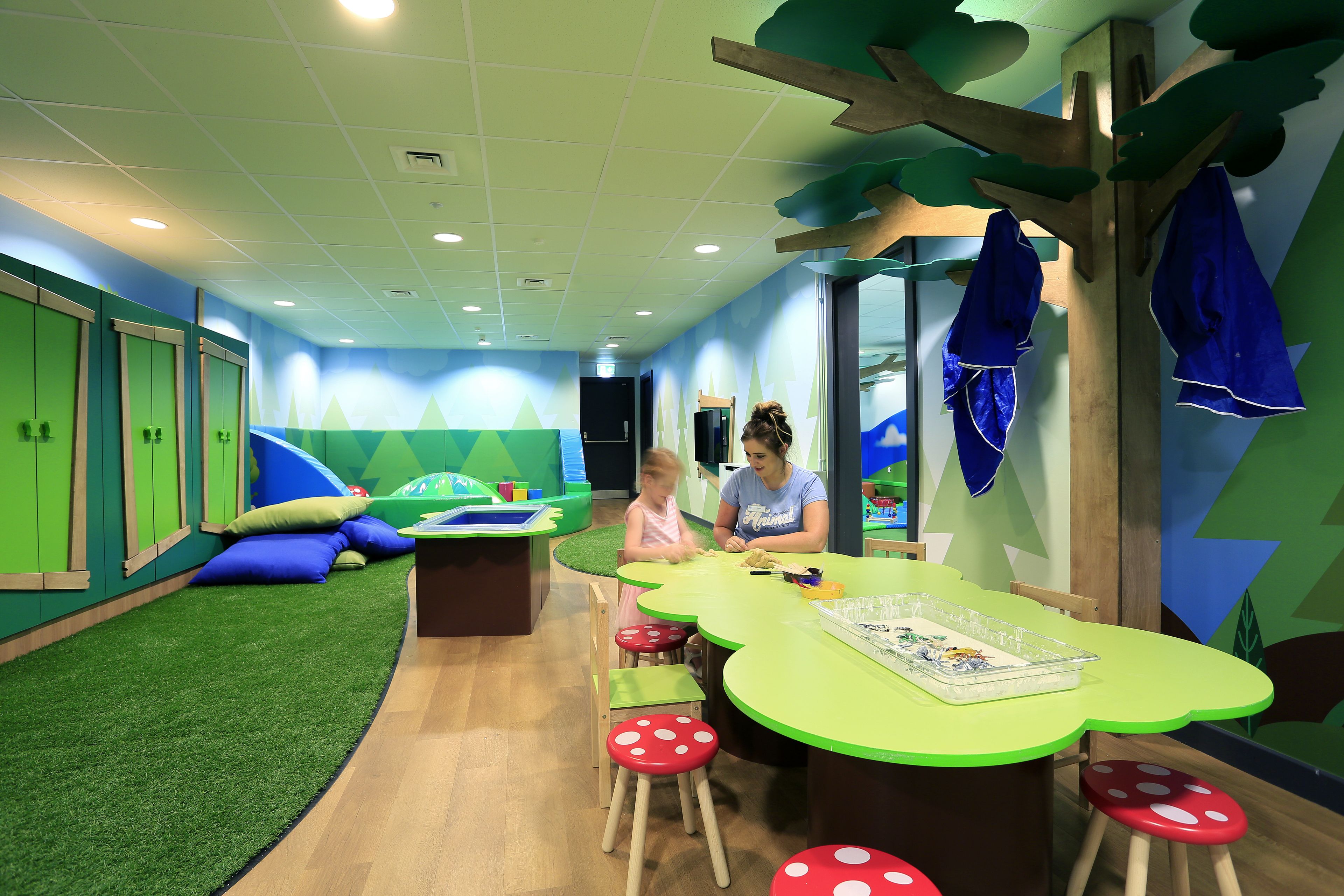 A children's play area with green and blue furniture, a table with a tray of snacks, and a mural of a mountain and tree.