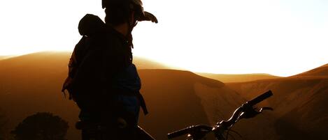 A person standing next to a bicycle with a backpack, overlooking a hilly landscape during sunset.