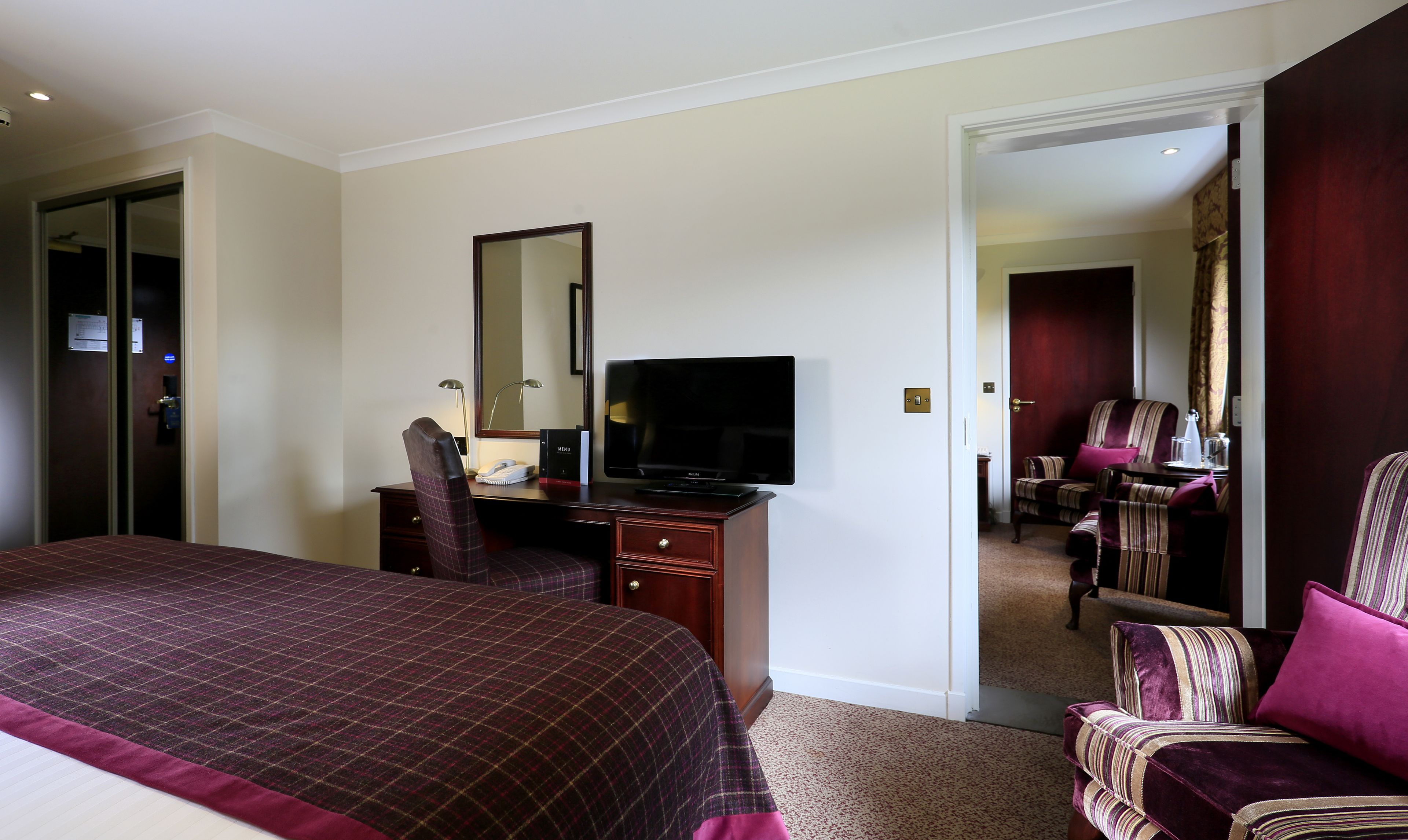 Ein Hotelzimmer mit einem großen Bett, einem Schreibtisch, einem Flachbildfernseher, einem Sessel und einem Spiegel.