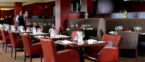 En restaurang med flera bord dukade för middag, röda stoppade stolar och en servitör i bakgrunden.