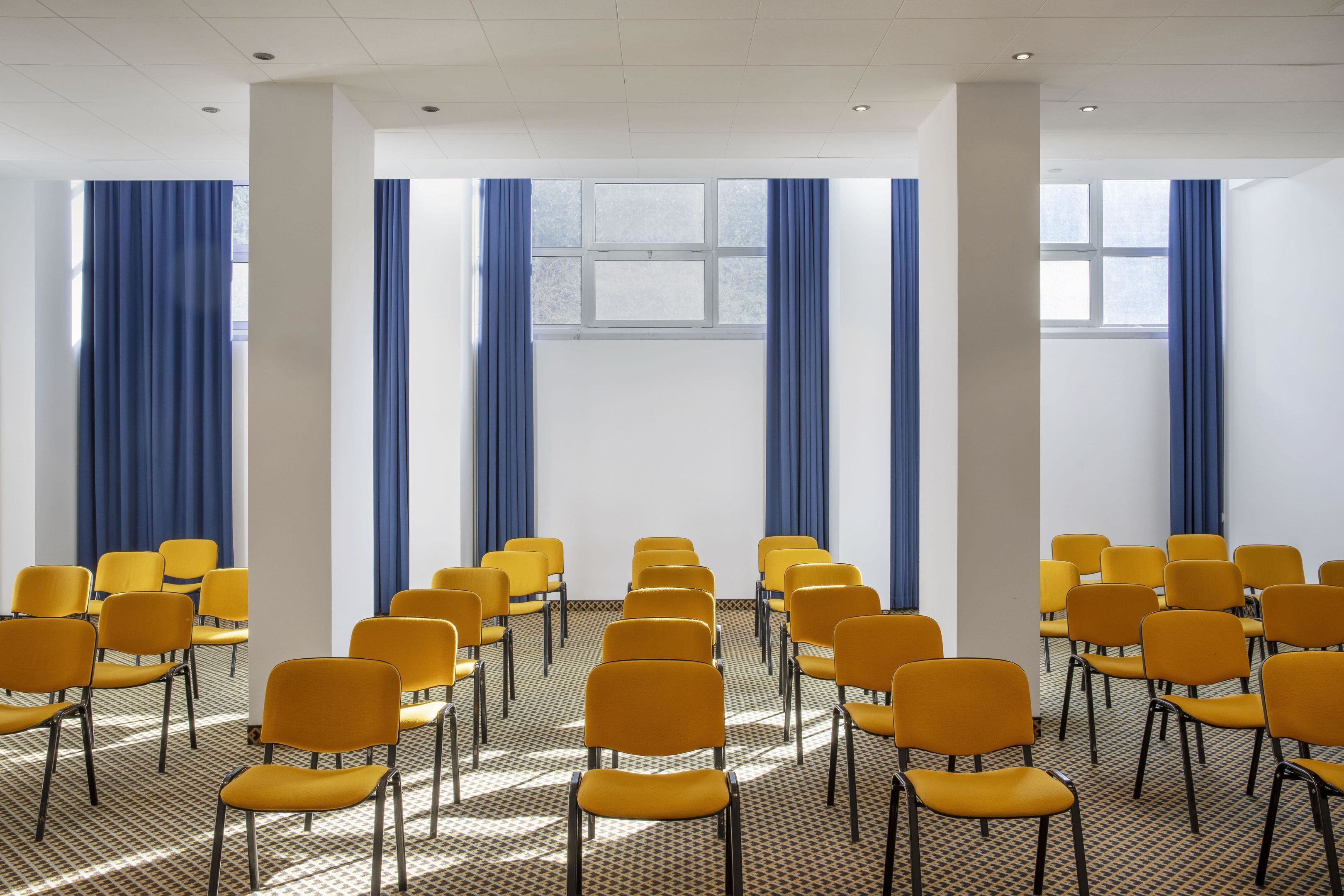 A conference room with rows of yellow chairs and blue curtains, large windows, and a patterned floor.