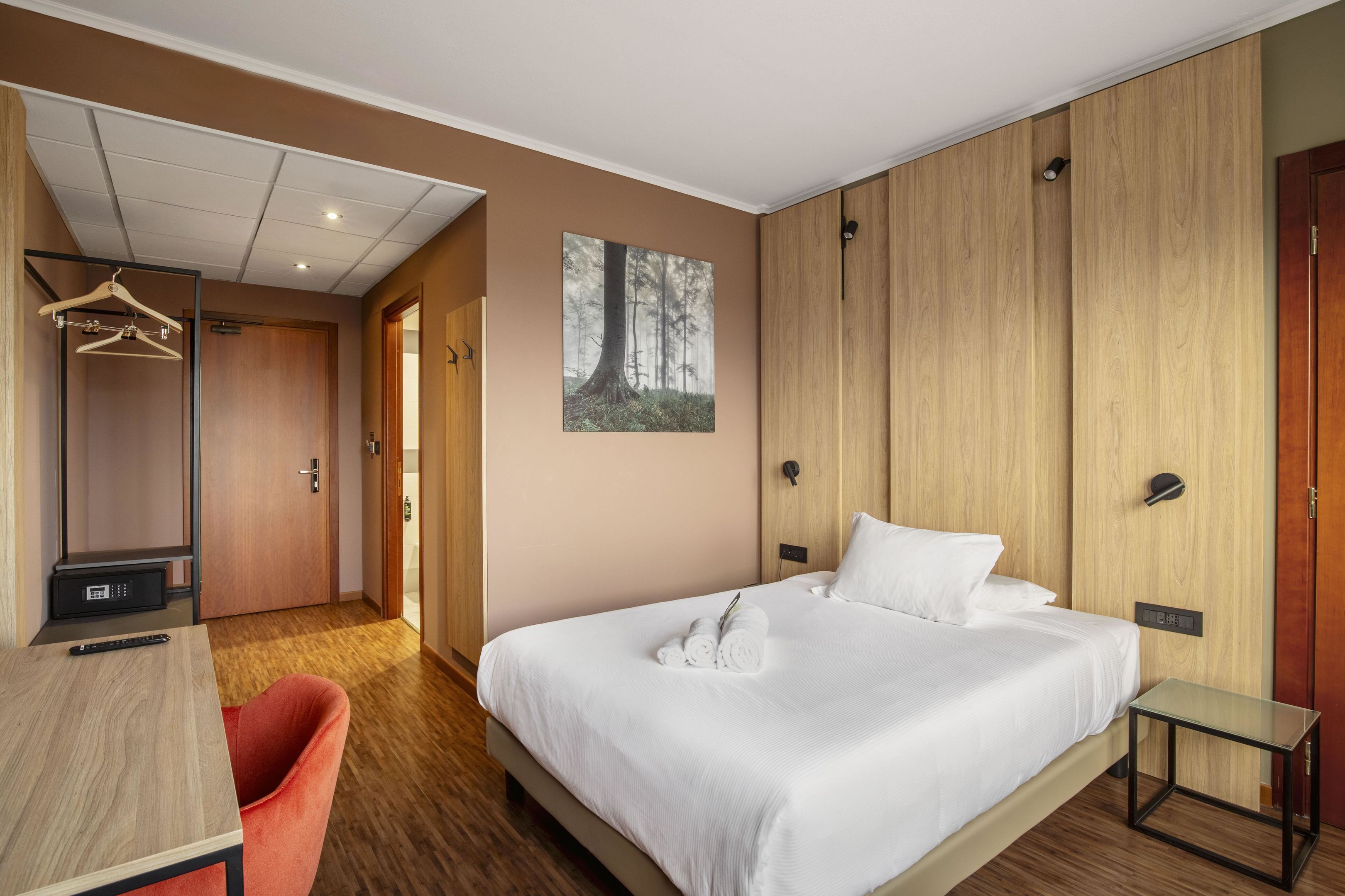 A hotel room with a bed, a desk, a chair, a wardrobe, and a painting of a forest.