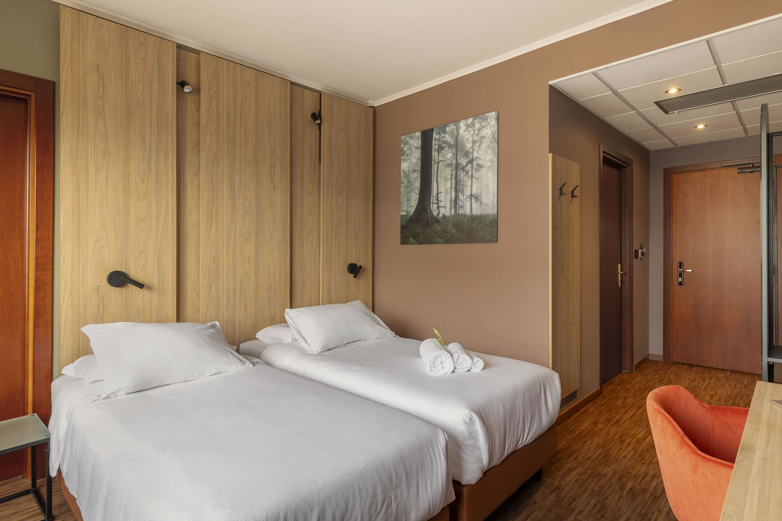 Ett hotellrum med två sängar, en garderob i trä, en målning som föreställer en skog och ett skrivbord i trä med en stol.