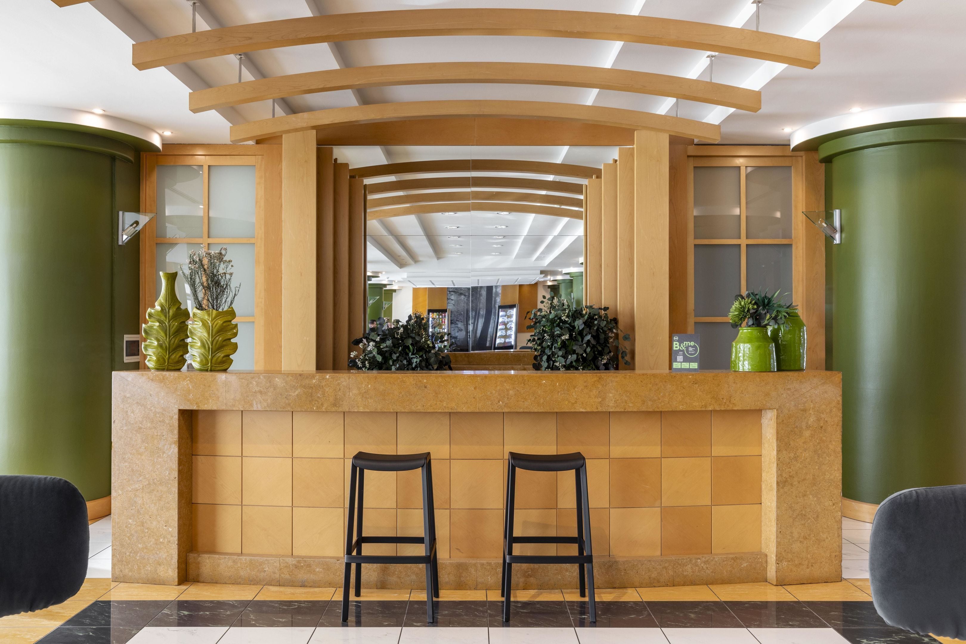 Un espace d’accueil doté d’un comptoir de réception en bois aux lignes courbes, de deux tabourets de bar et d’une grande fenêtre donnant sur l’intérieur.
