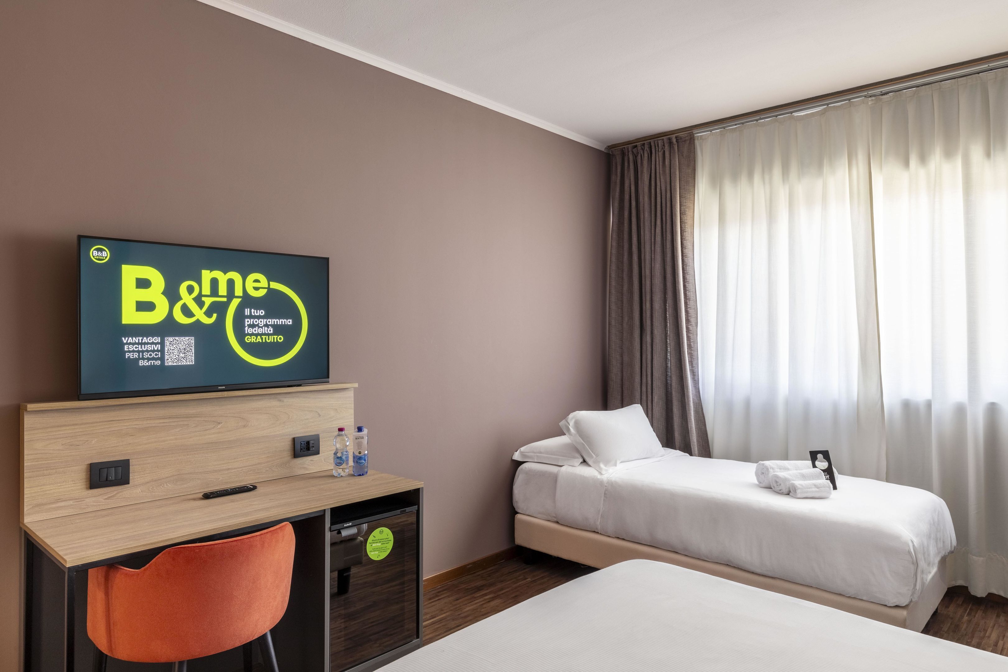 Habitación de hotel con cama, escritorio con silla, televisor mostrando un programa y ventana con cortinas.