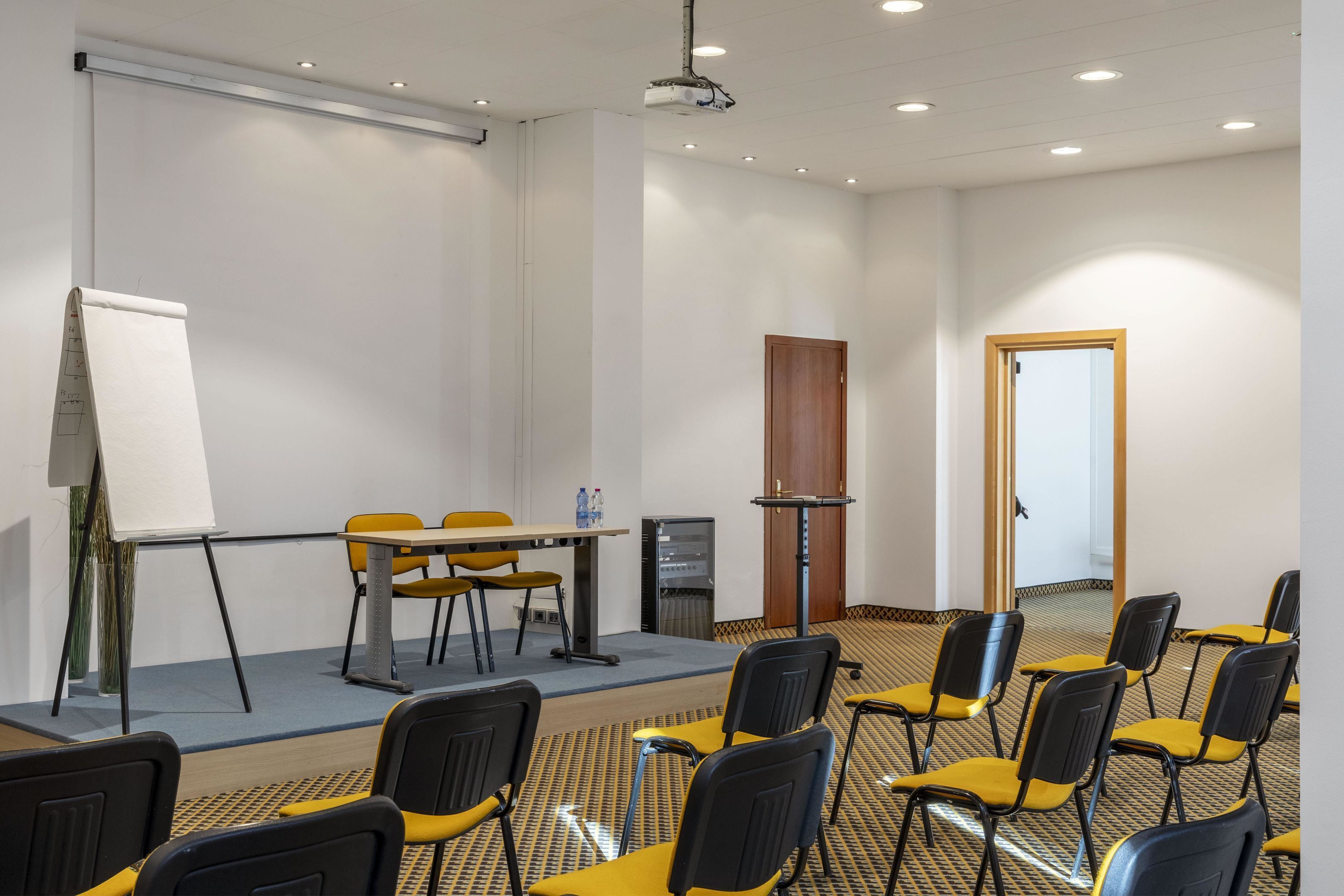 Uma sala de conferências com quadro branco, projetor, mesa e cadeiras, e piso coberto de carpete.