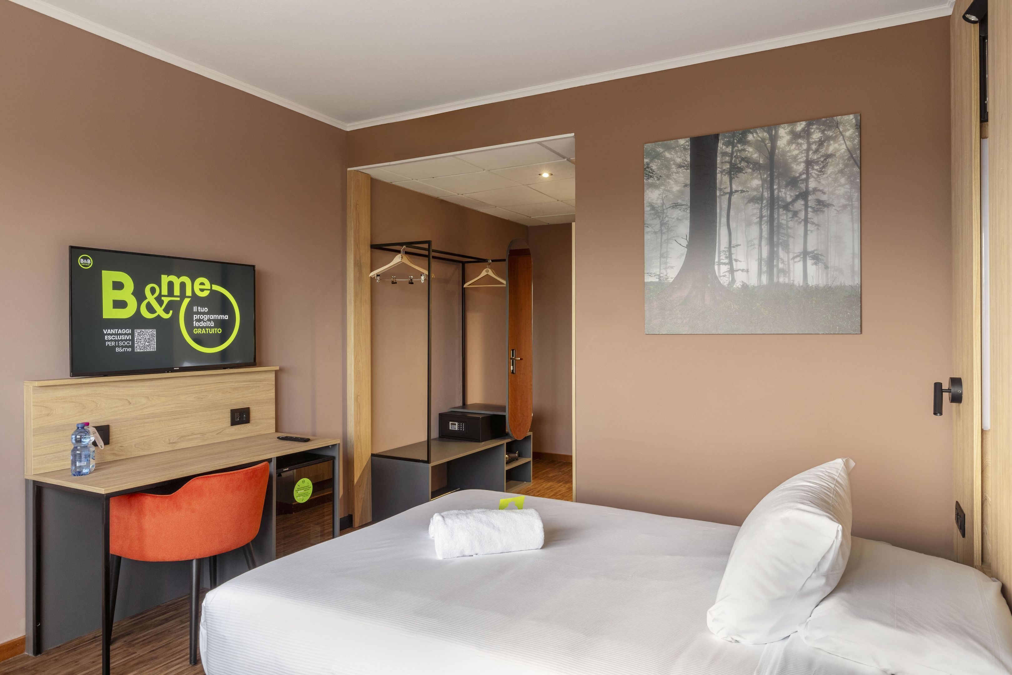 A hotel room with a bed, a desk with a chair, a coat rack, and a painting on the wall.