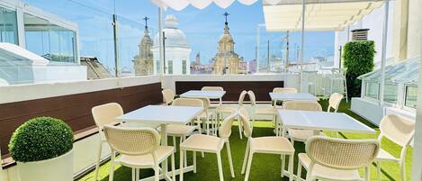 A rooftop terrace with white tables and chairs, a white umbrella, and a view of city buildings.