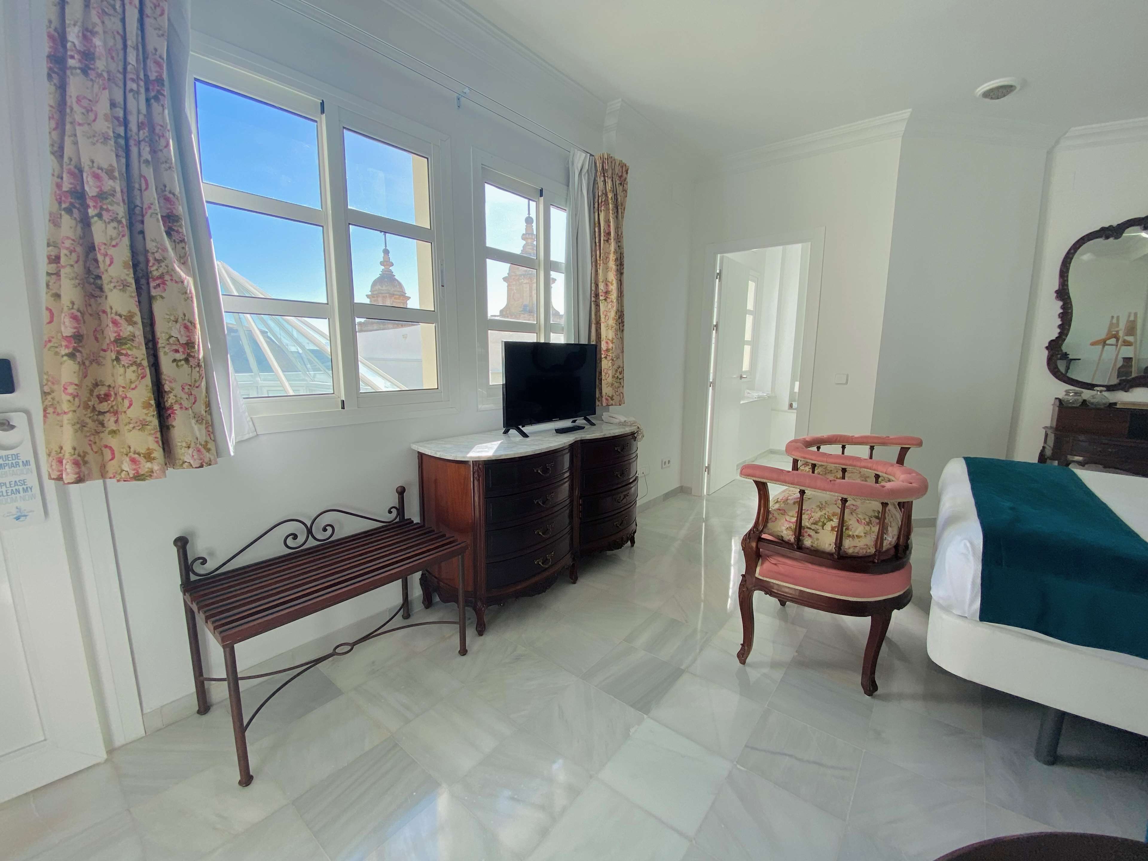 A hotel room with a bed, a television on a wooden dresser, a bench, a chair, and large windows with curtains.