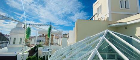 Una terraza en la azotea con techo de cristal, mobiliario blanco y un edificio de fondo.