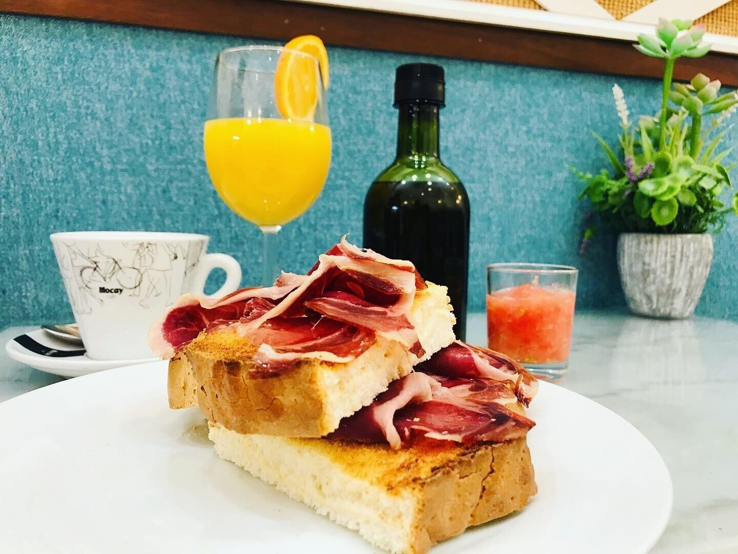 A breakfast plate with toast and ham, a glass of orange juice, a bottle of olive oil, a cup with a horse design, and a small potted plant.