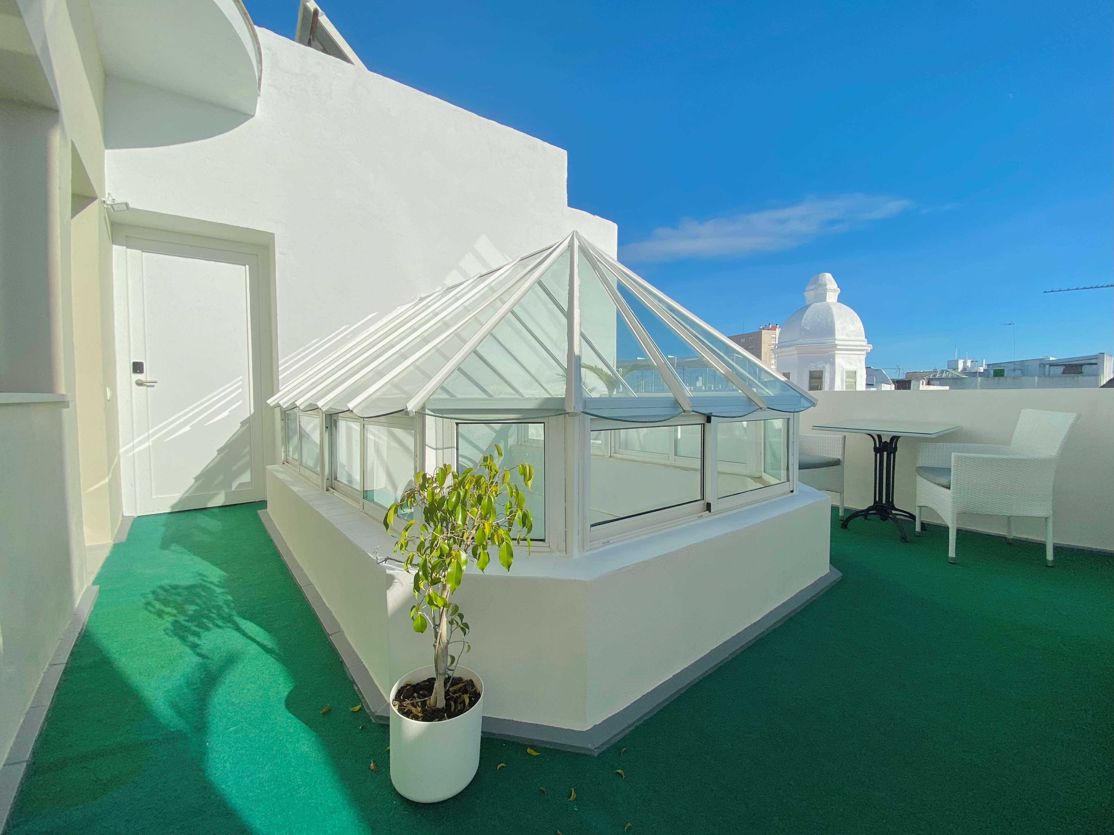 Una terraza en la azotea con cerramiento de vidrio, una planta en maceta y una mesa blanca con sillas.