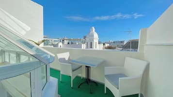 A rooftop terrace with white wicker furniture, a glass table, and a view of a white building with a dome in the background.