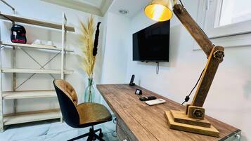 A modern office desk with a wooden finish, a leather chair, a desk lamp, a vase with dried plants, a shelf with items, and a mounted TV.