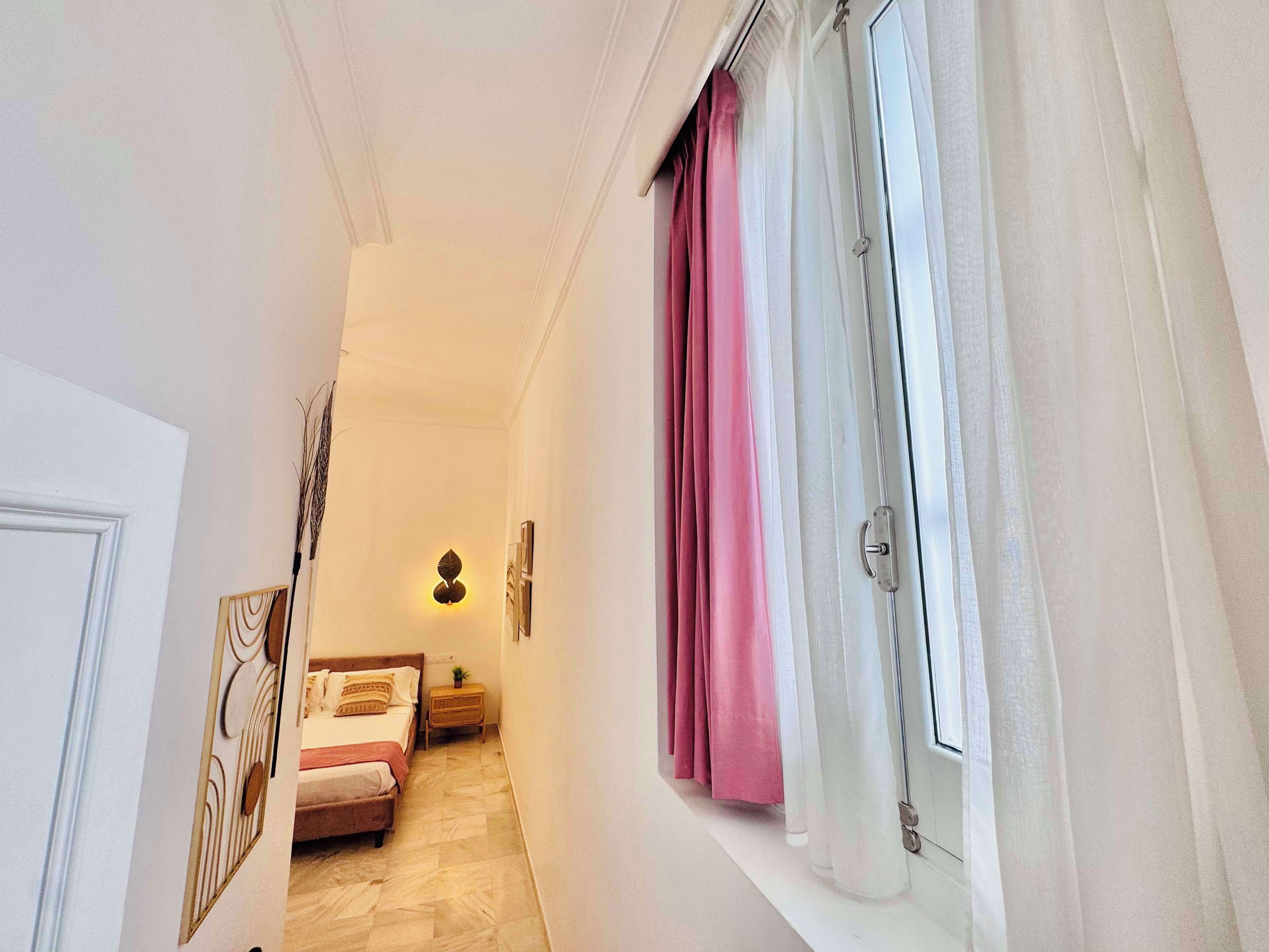 A narrow hallway leading to a bedroom with a bed, a nightstand, and a wall-mounted light fixture.