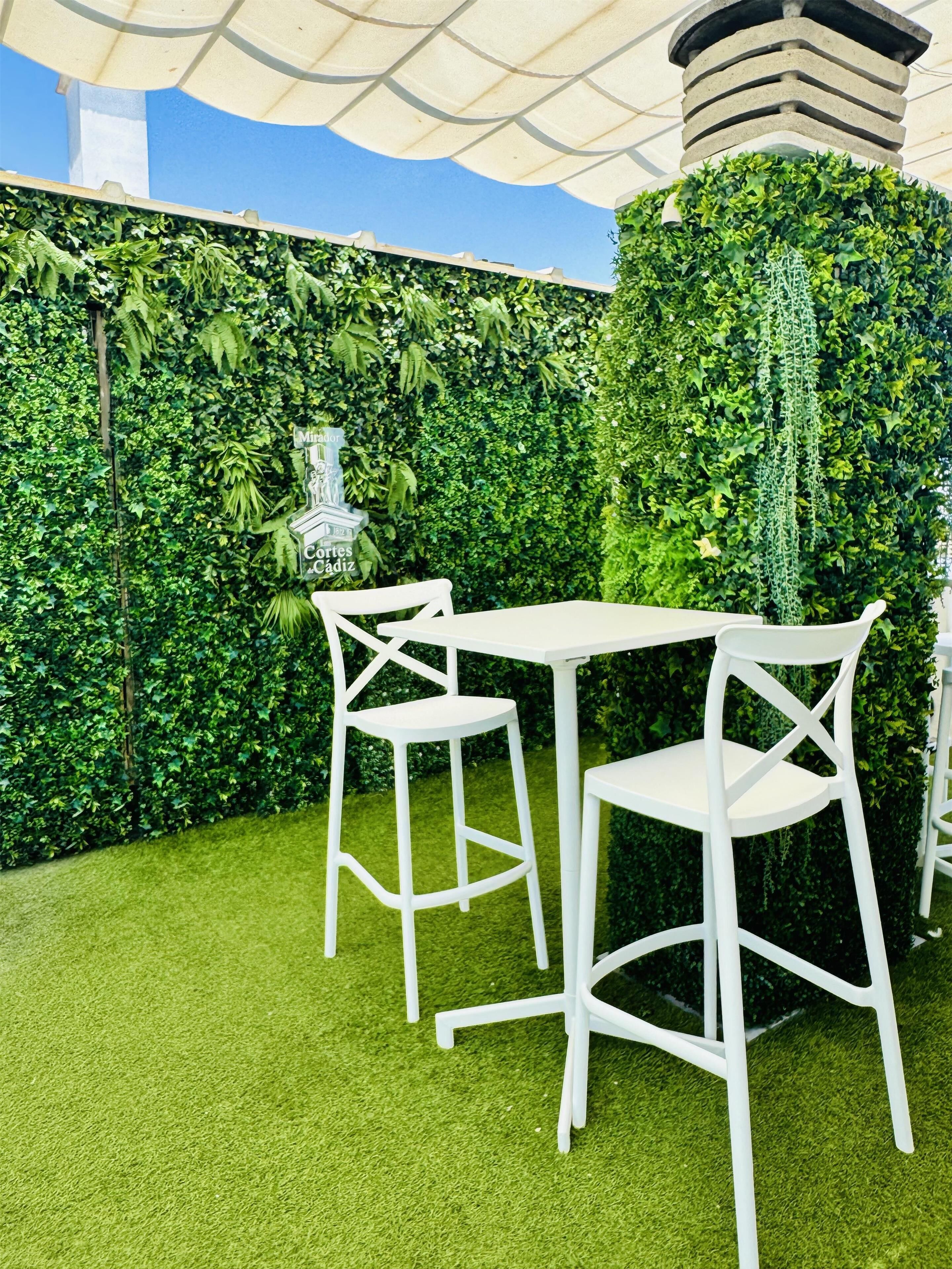 Un patio con sillas y una mesa blancos, rodeado de exuberante follaje verde y una pared decorativa.