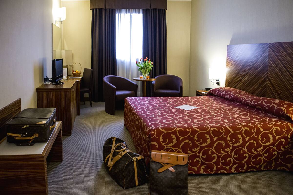 Une chambre d’hôtel avec un grand lit, deux fauteuils, un bureau avec un ordinateur, une petite table avec un vase de fleurs et une valise posée sur le sol.