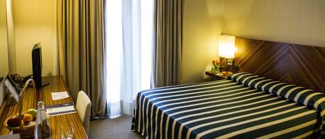 Habitación de hotel con una cama grande, un escritorio con computadora, una lámpara y un ventanal con cortinas.