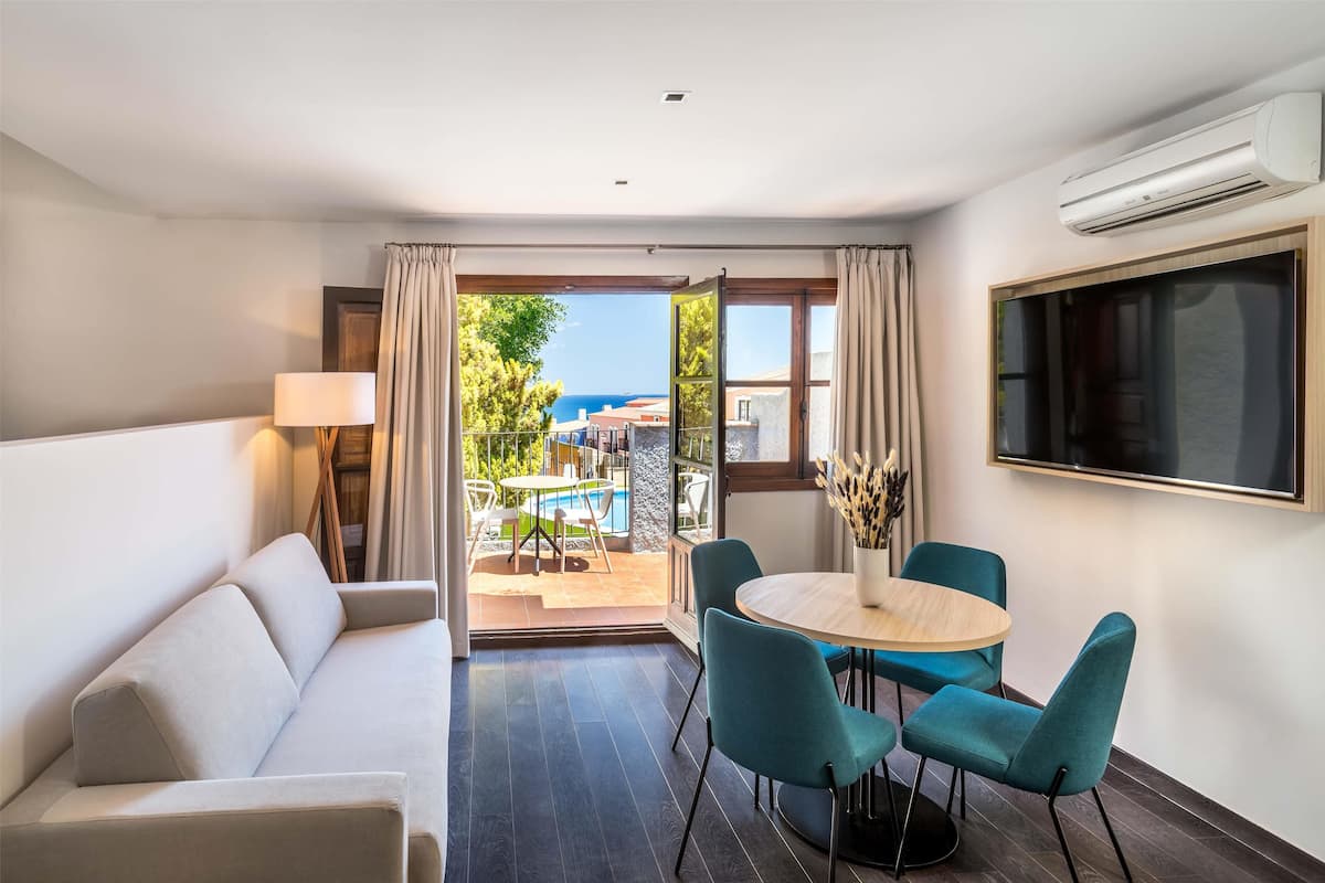 Une chambre moderne avec un canapé, une table ronde entourée de chaises, un téléviseur et une vue sur la mer grâce à de grandes fenêtres.