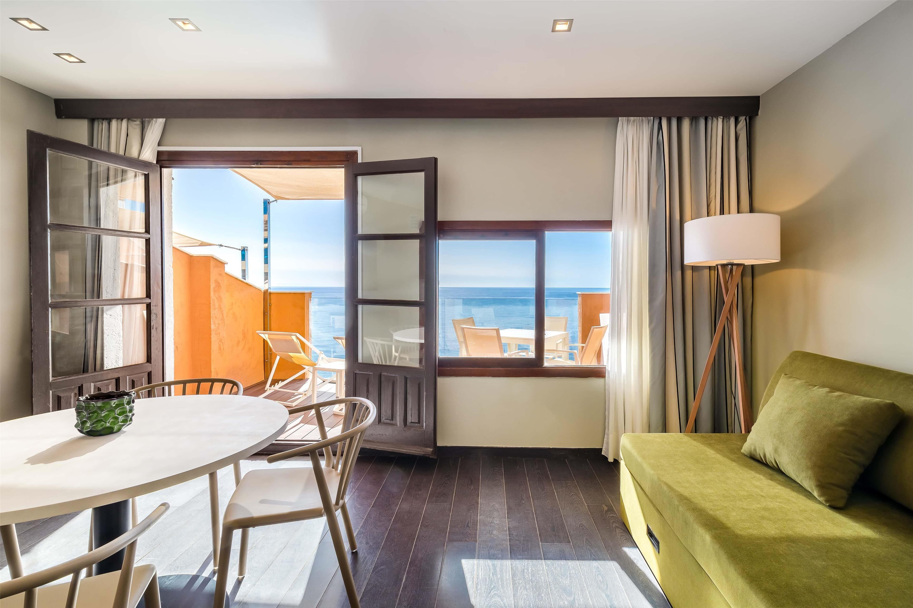 A room with a green sofa, a dining table with chairs, a view of the sea, and a large window.