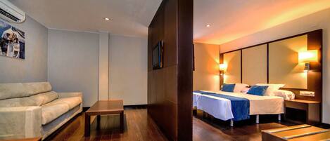 A hotel room with a sofa, a small table, a bed with two pillows, a nightstand, and a wall-mounted TV.