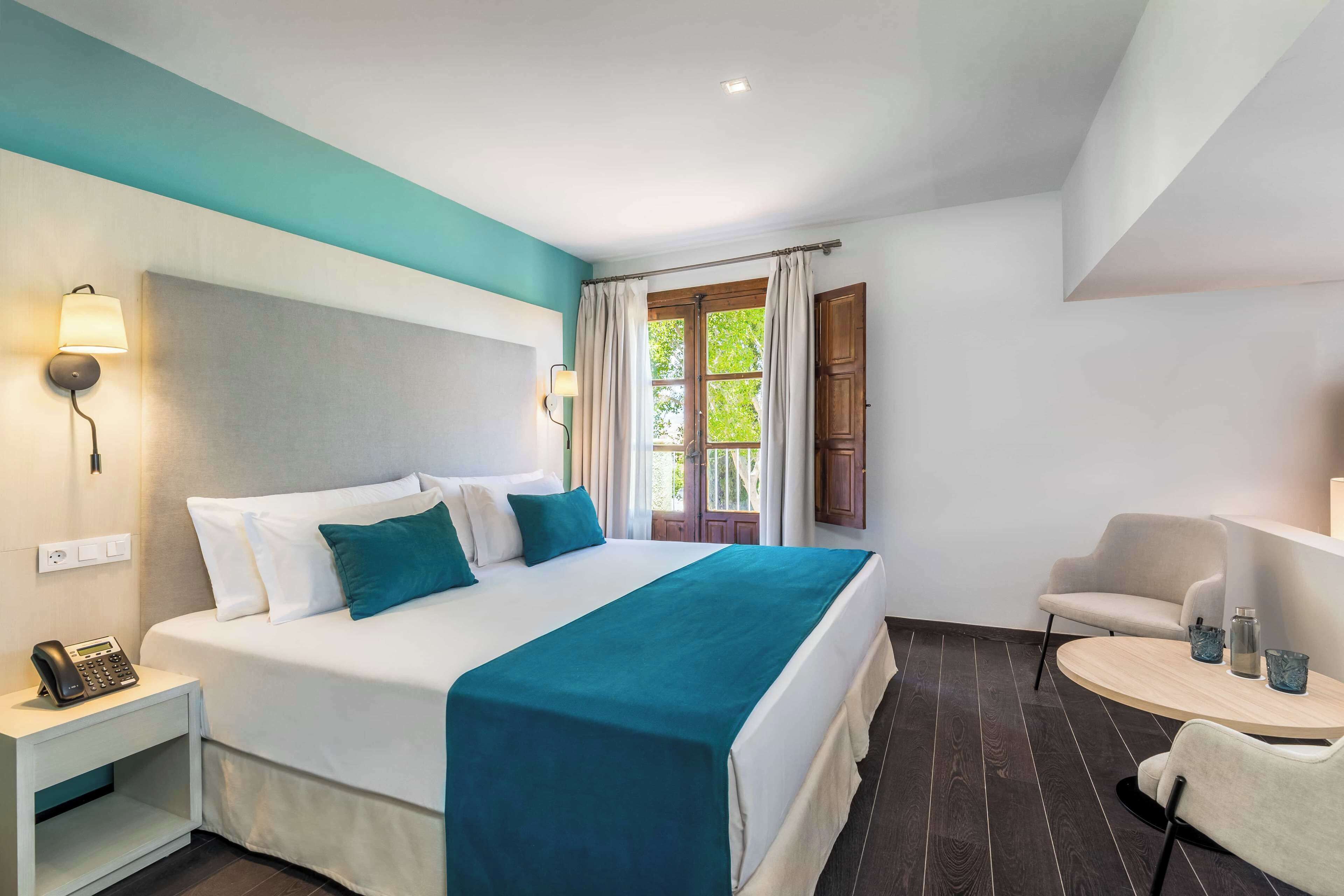 Habitació d'hotel amb un llit, una taula de nit amb un telèfon, una cadira, una taula petita amb gots i una finestra amb cortines.