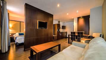 Una habitación de hotel moderna con una cama grande, un sofá, un televisor montado en la pared, una zona de comedor con mesa y sillas, y una kitchenette.