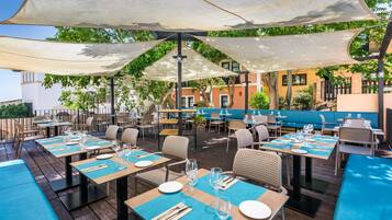 An outdoor dining area with tables and chairs, shaded by large umbrellas, and a view of buildings in the background.