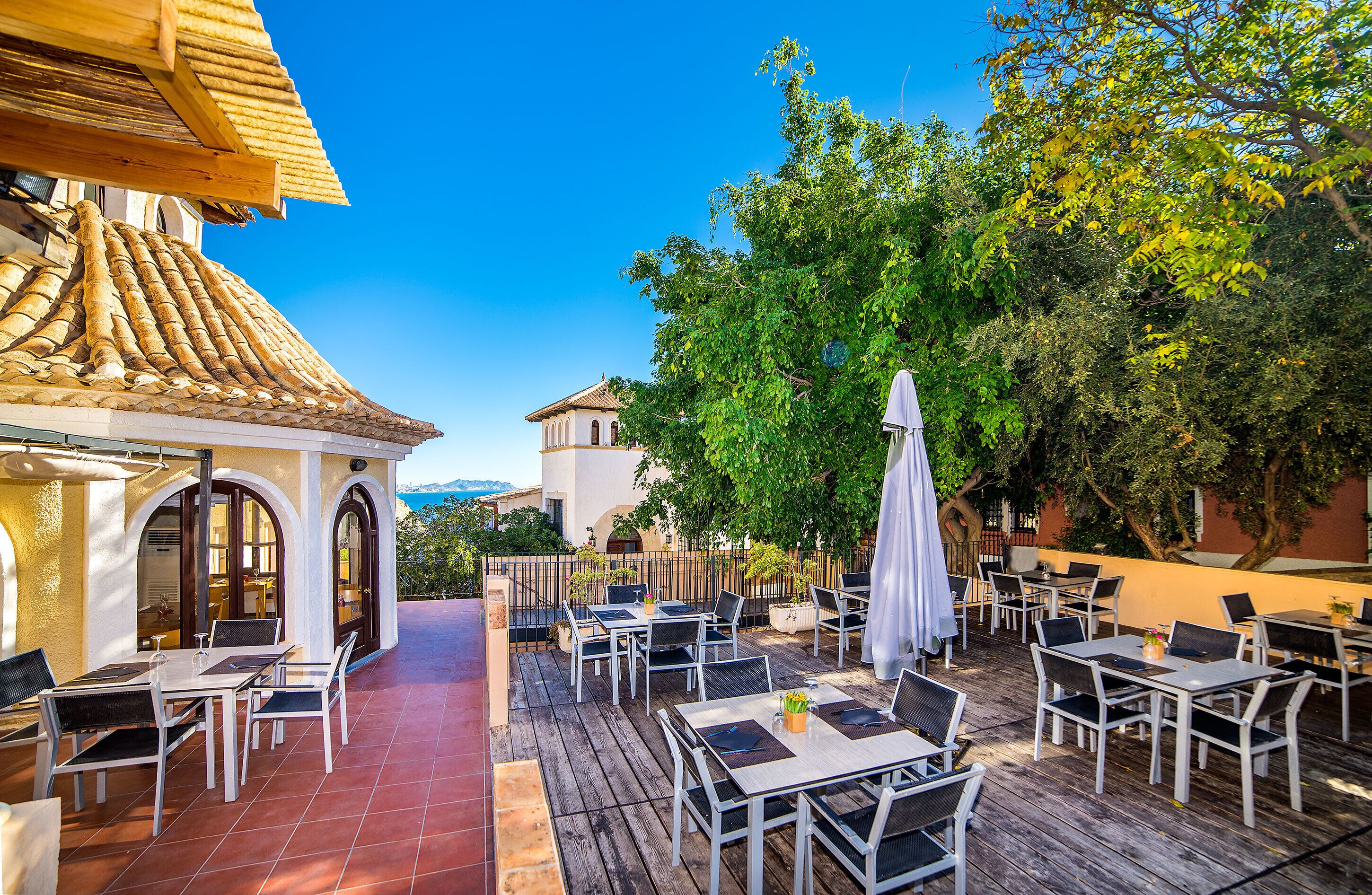 An outdoor patio with tables and chairs, a covered seating area, and a view of a building with a tiled roof.