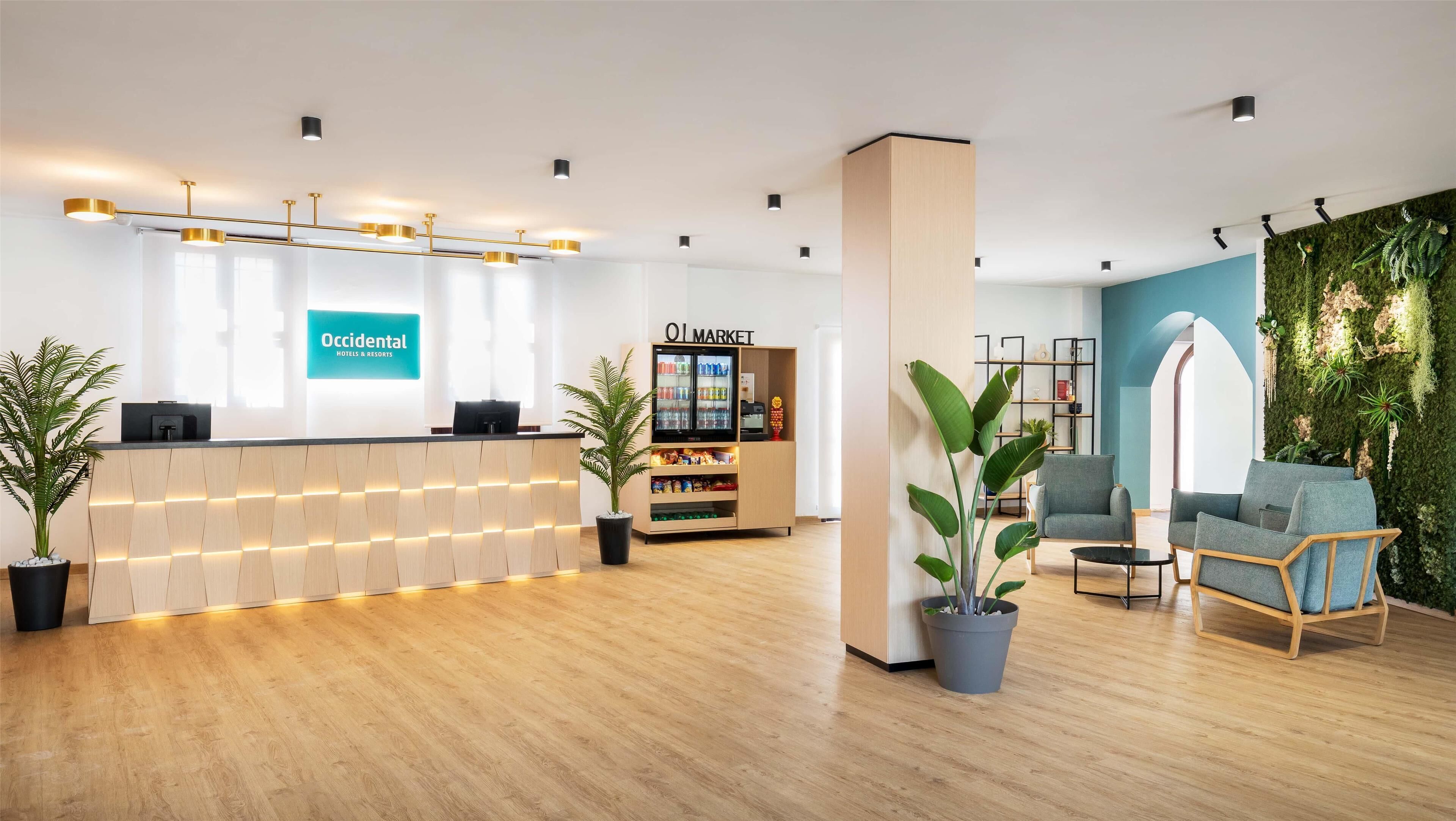 A hotel reception area with a wooden reception desk, a 'Market' refrigerator, seating area with chairs, and a green wall with plants.