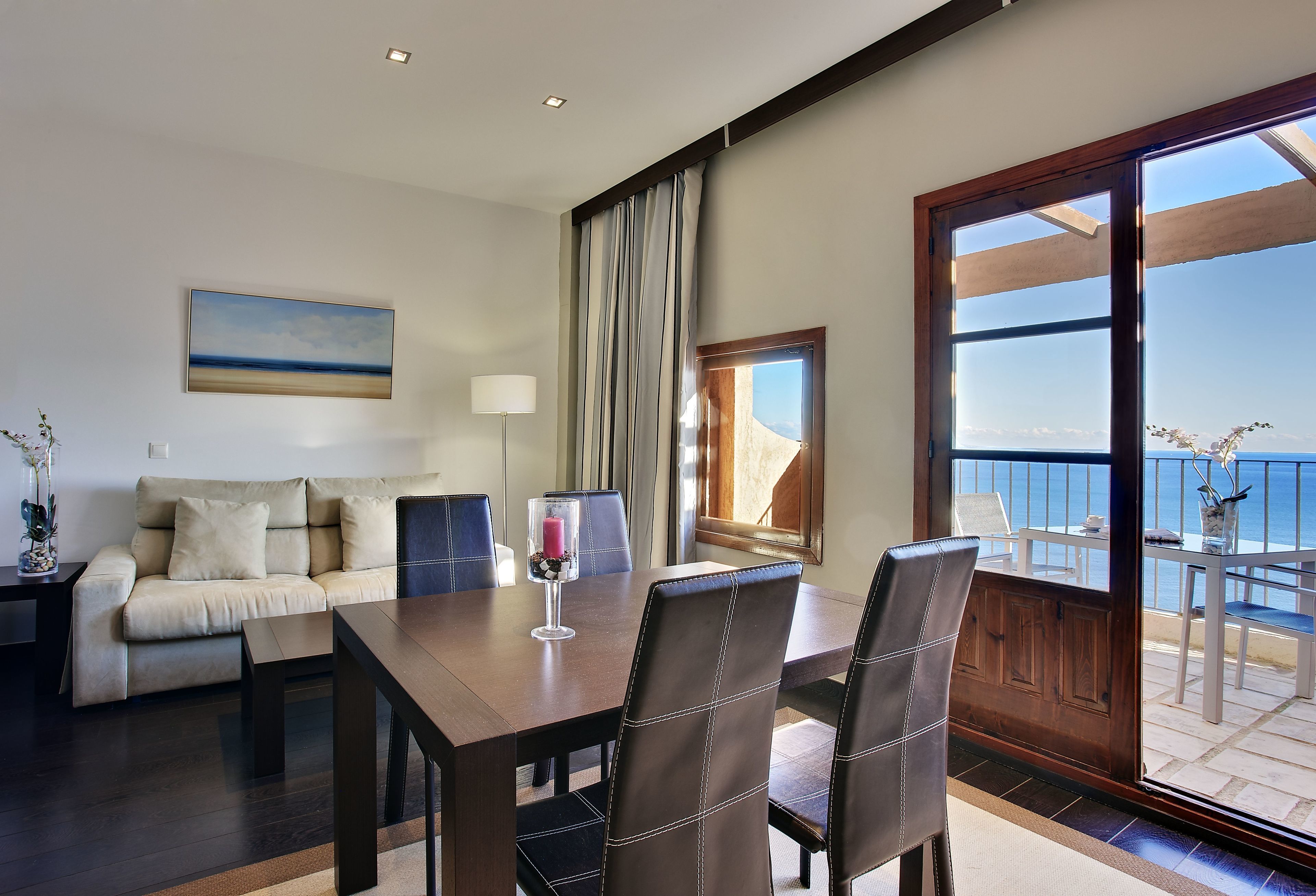A modern living room with a dining area, a sofa, a coffee table, and a view of the sea through a large window.