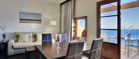 A modern living room with a dining area, a sofa, a coffee table, and a view of the sea through a large window.