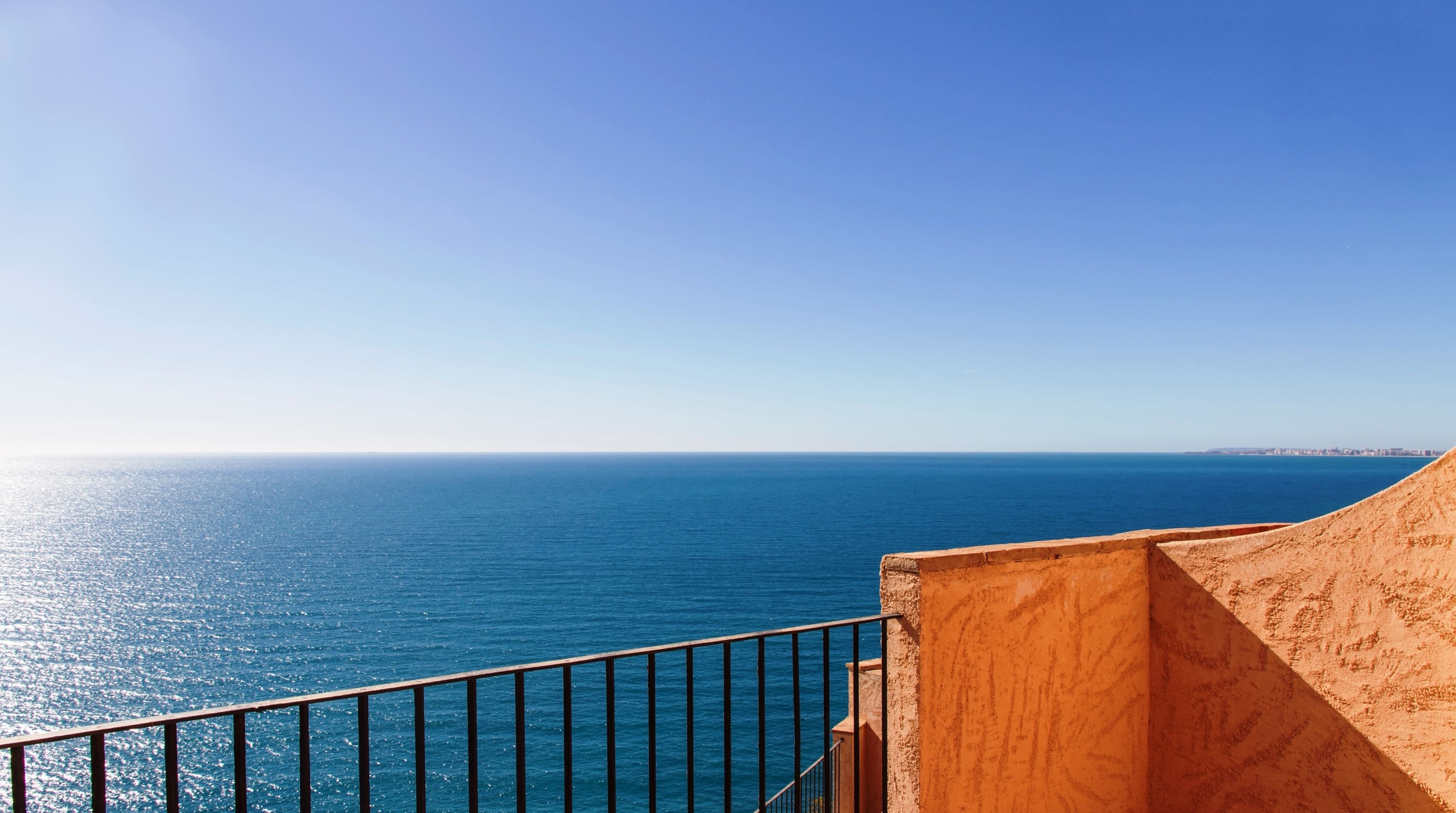 Een kustgezicht met heldere lucht, een balkon met metalen hekwerk en een zandstrand.