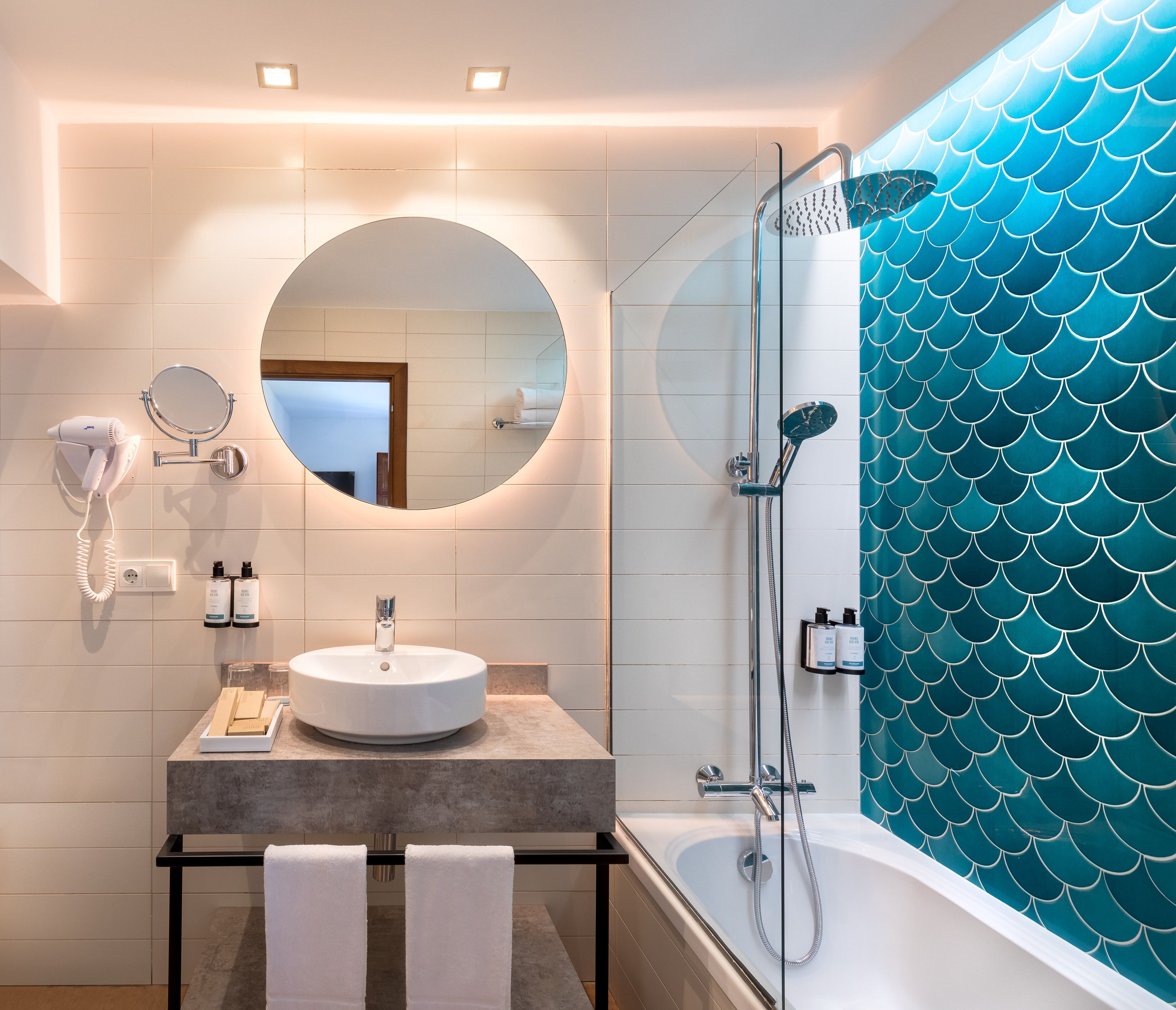 Un baño moderno con un espejo redondo, un secador de pelo, un lavabo y una ducha con un patrón de escamas color turquesa en la pared.