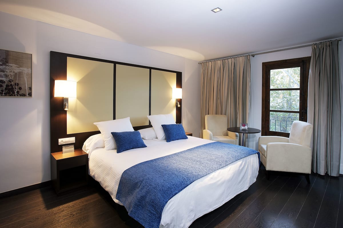 Une chambre d’hôtel avec un grand lit, deux lampes de chevet, une chaise, une petite table et une fenêtre avec des rideaux.
