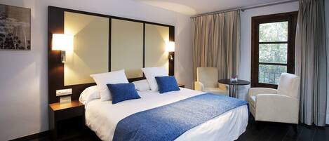 Habitación de hotel con una cama grande, dos lámparas de noche, una silla, una mesita y una ventana con cortinas.