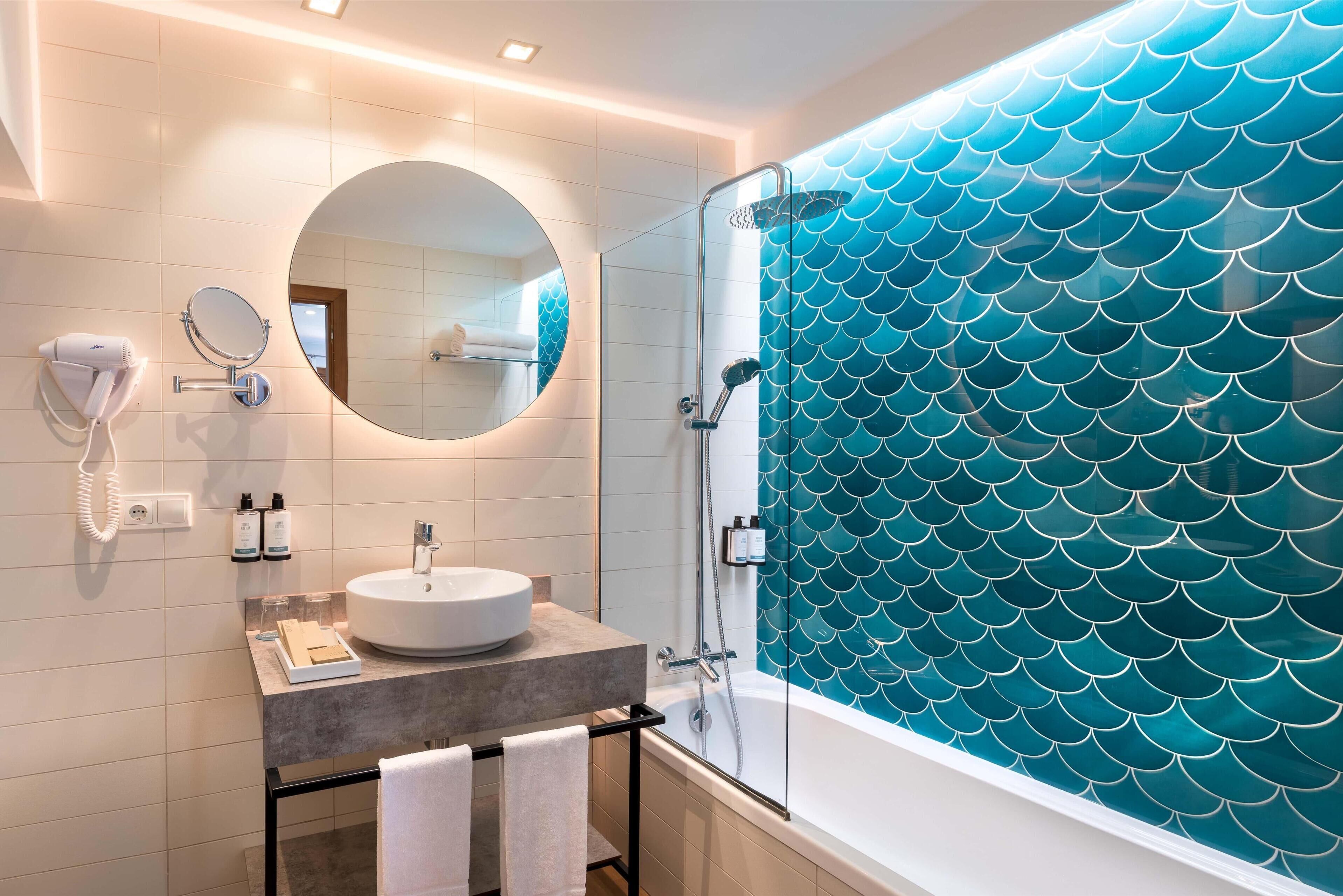 Un baño moderno con un espejo redondo, un secador de pelo, un lavabo y una ducha con cerramiento de vidrio y un revestimiento de azulejos tipo escama de pez azul.