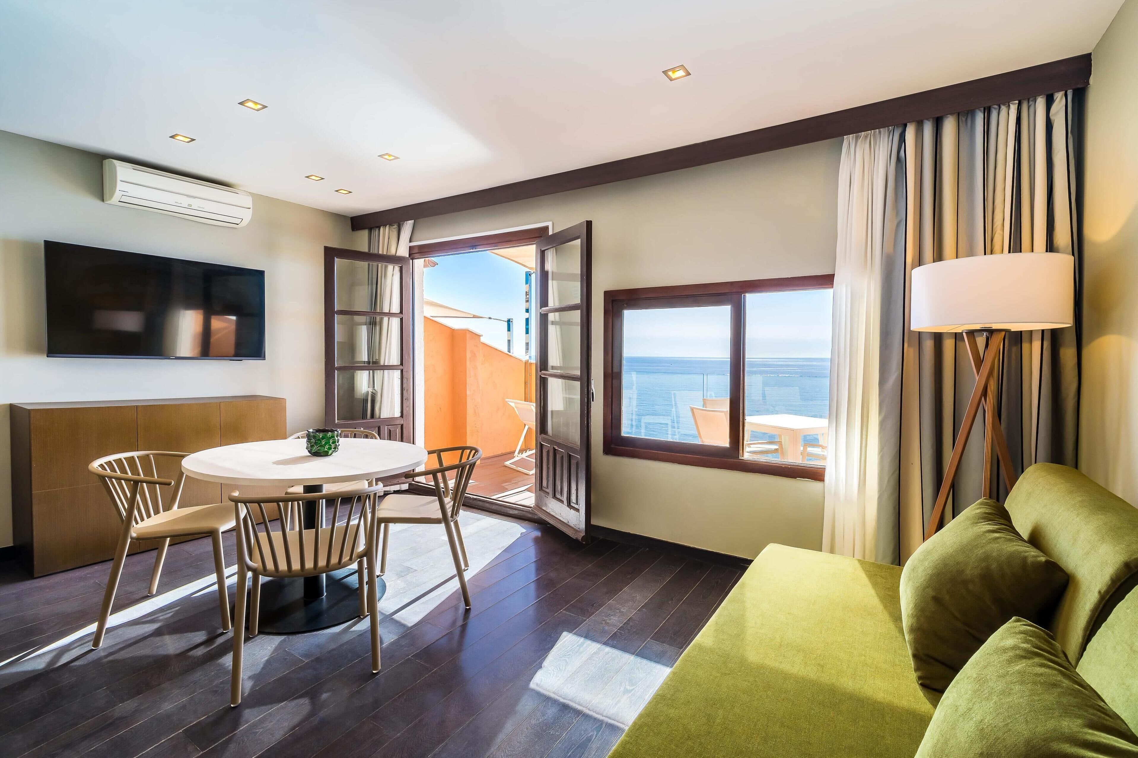 A hotel room with a dining area, a sofa, a TV, and a large window with curtains.