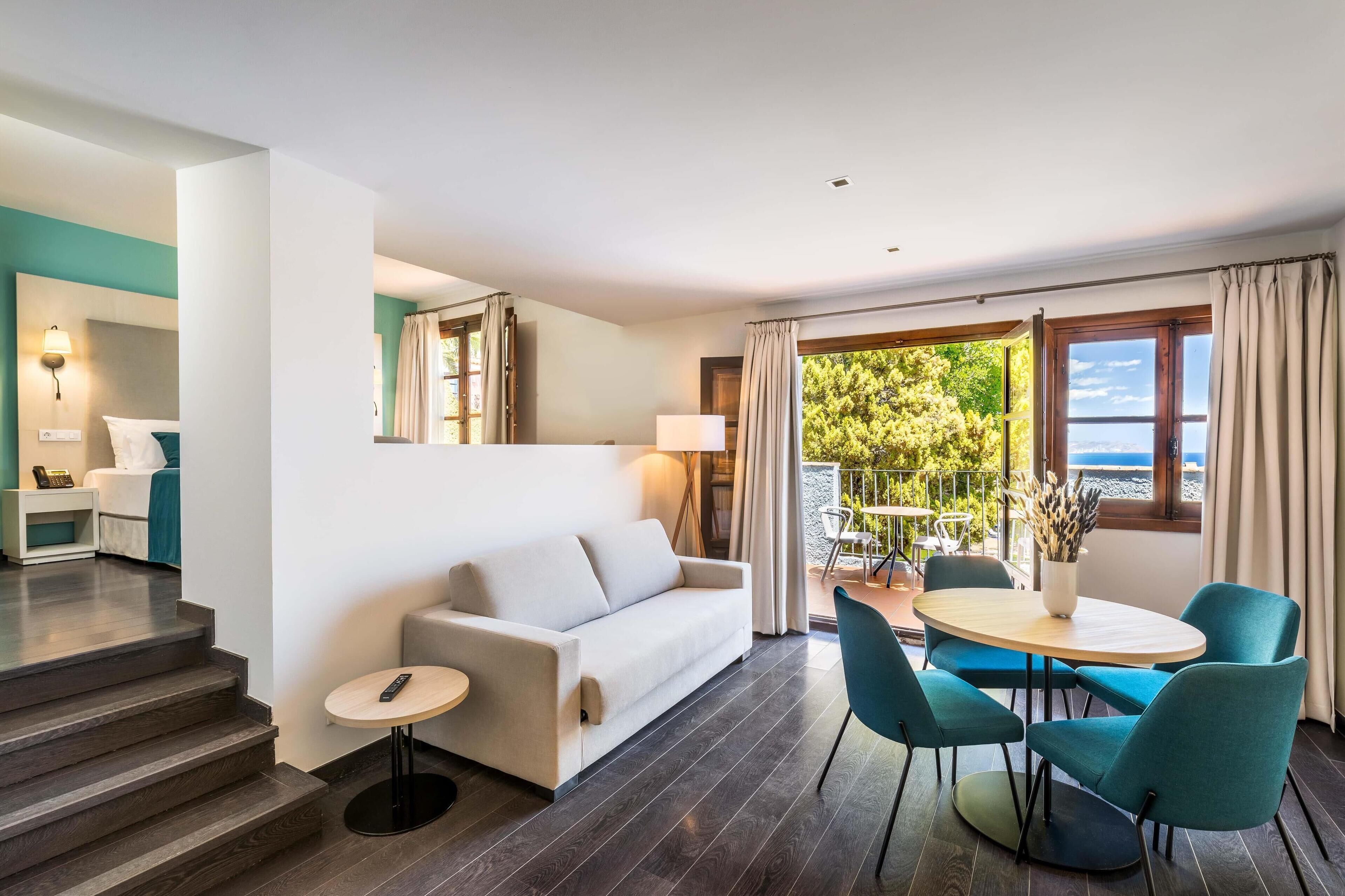 A modern hotel room with a sofa, a small table, and a balcony with a view of the sea.