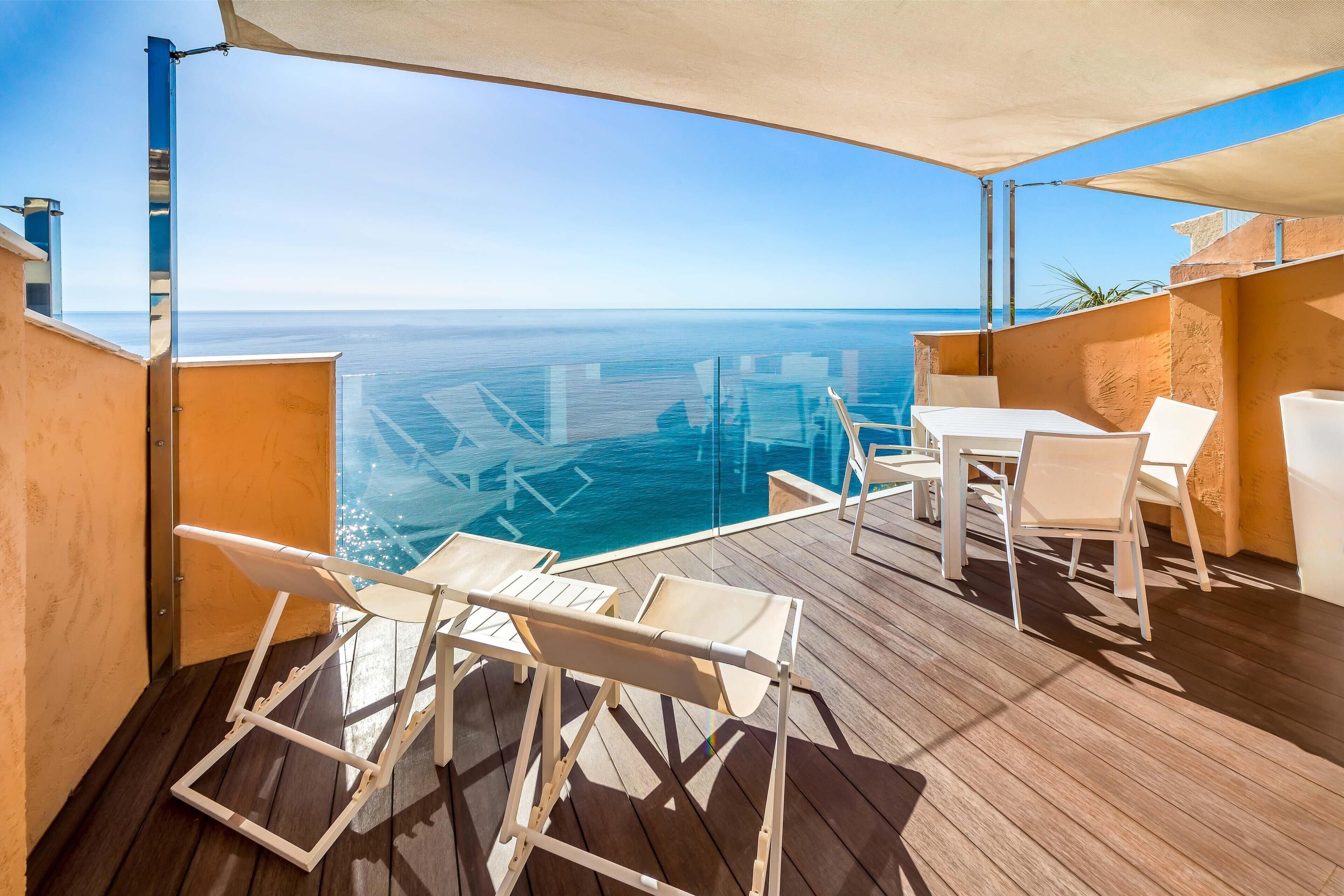 A balcony with a view of the sea, wooden flooring, white chairs, and a table.