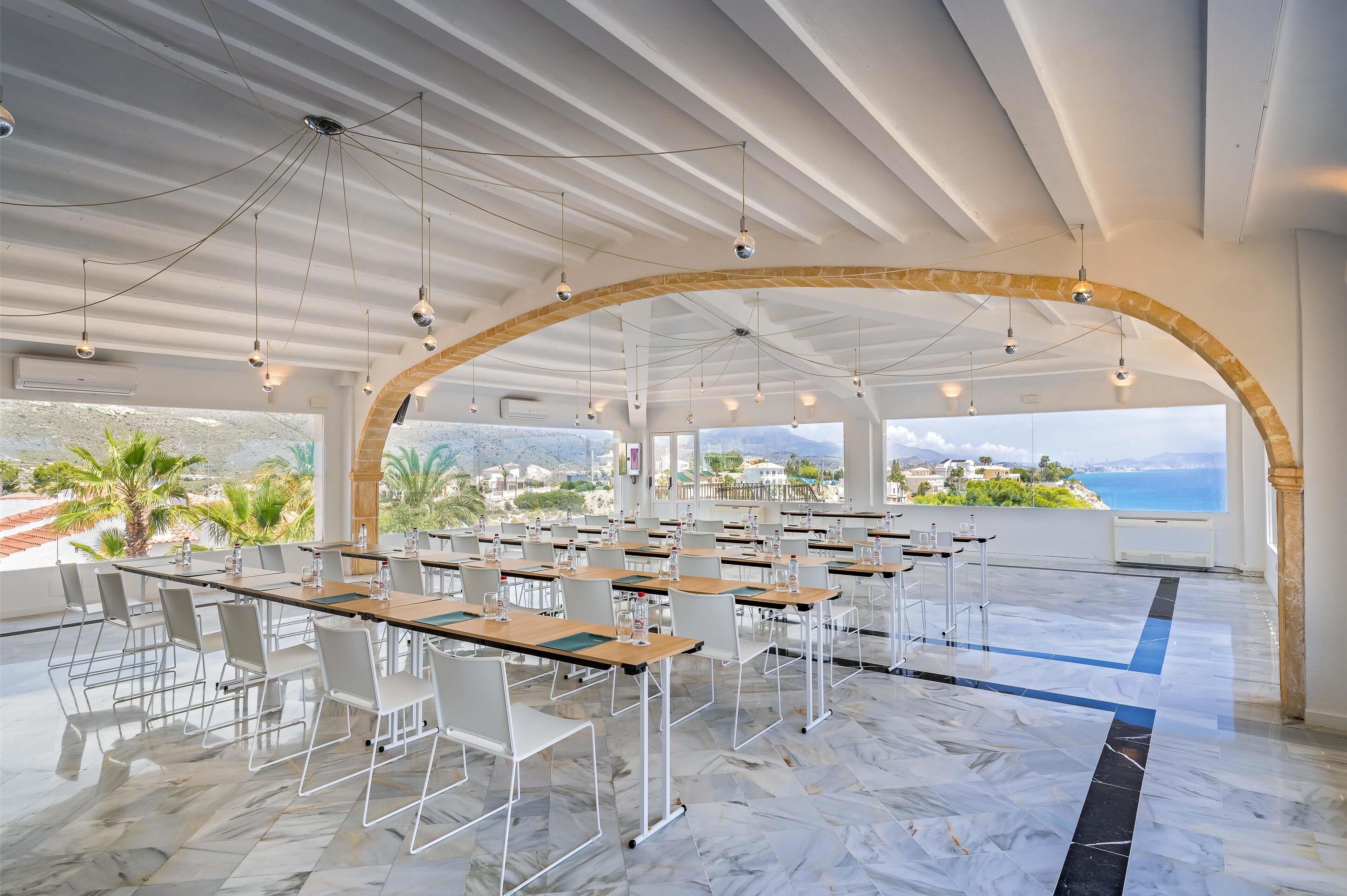 A spacious conference room with marble flooring, white chairs, and tables, featuring a large arched window overlooking a scenic landscape.