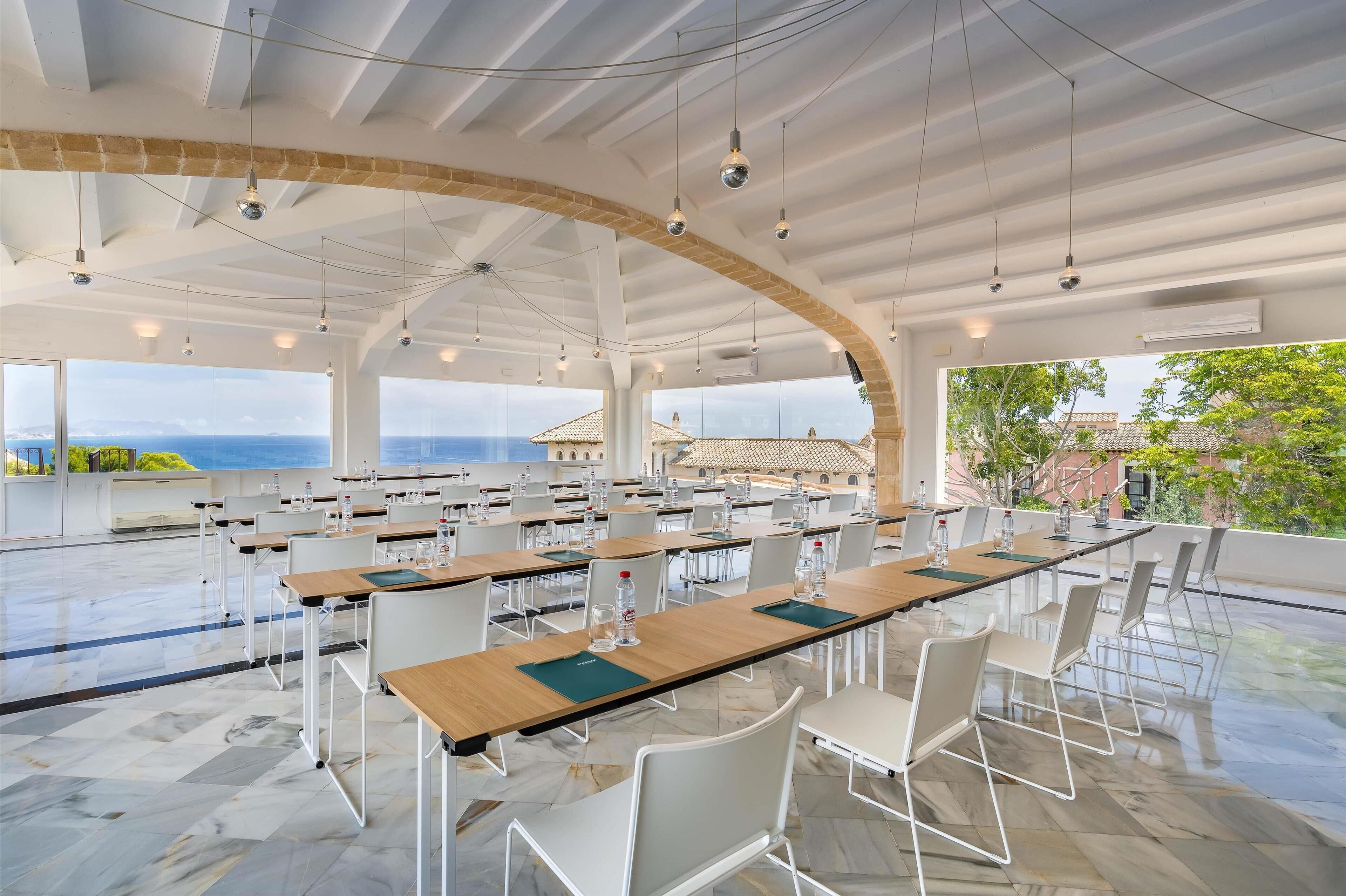 Un espacio interior amplio con mesas y sillas largas, ventanas grandes que ofrecen vistas al mar y edificios, y un techo alto con vigas a la vista y luces colgantes.