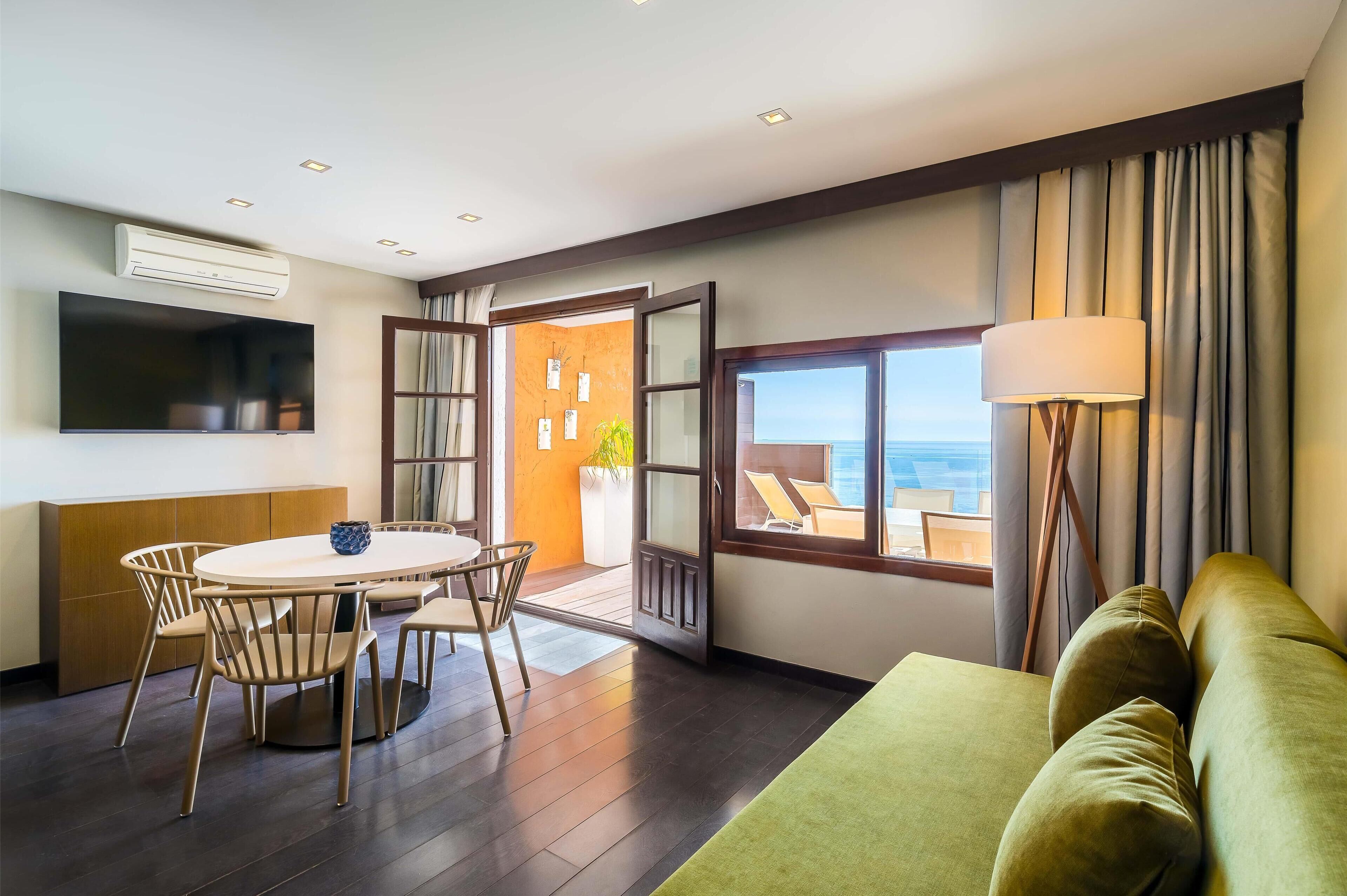 Habitación de hotel moderna con zona de comedor, sofá, televisor y un ventanal con vistas.