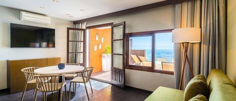 Habitación de hotel moderna con zona de comedor, sofá, televisor y un ventanal con vistas.