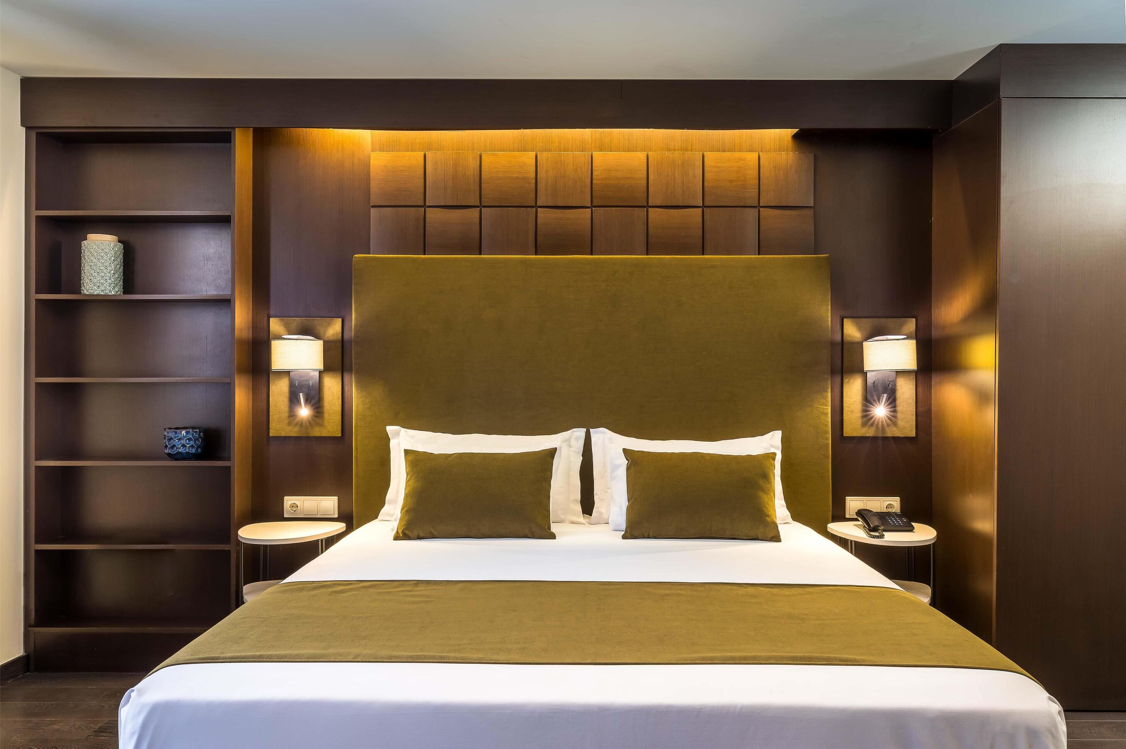 A modern hotel room with a large bed, two bedside tables, a small round table, and built-in shelving.