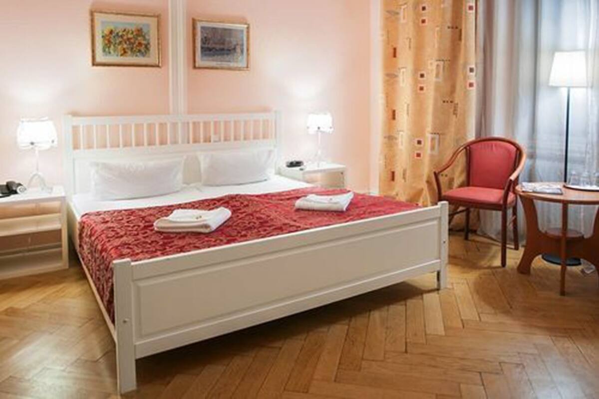 Ein ordentlich bezogenes Bett mit einer roten Decke, ein Holzstuhl, ein kleiner Tisch und eine Lampe.